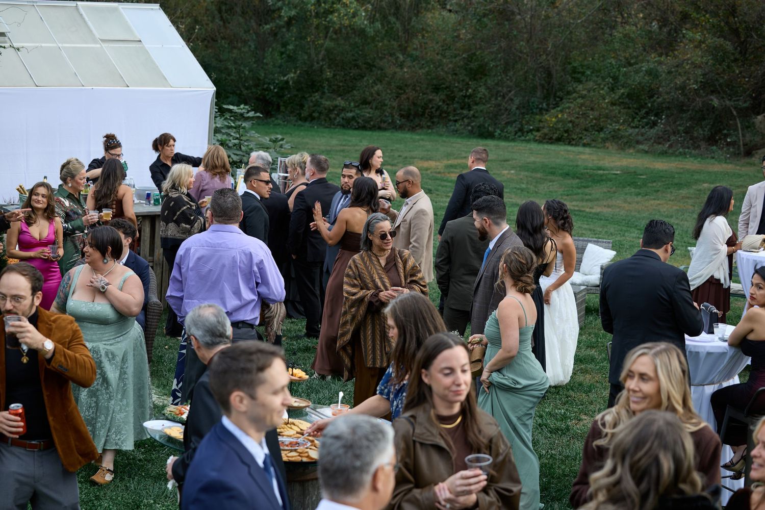 Wedding guests in formal attire mingle and chat on a grassy lawn near a white tent during an outdoor cocktail hour.