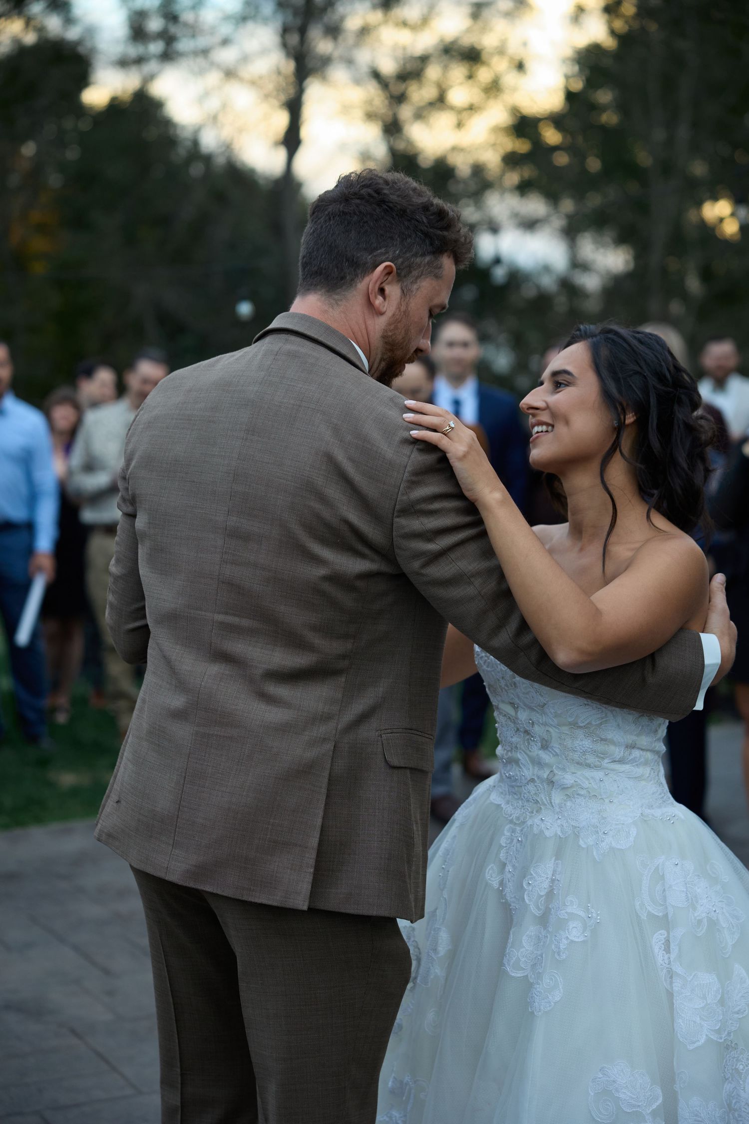 A bride in a lace wedding gown smiles and dances with her groom in a tweed suit as guests look on outdoors.
