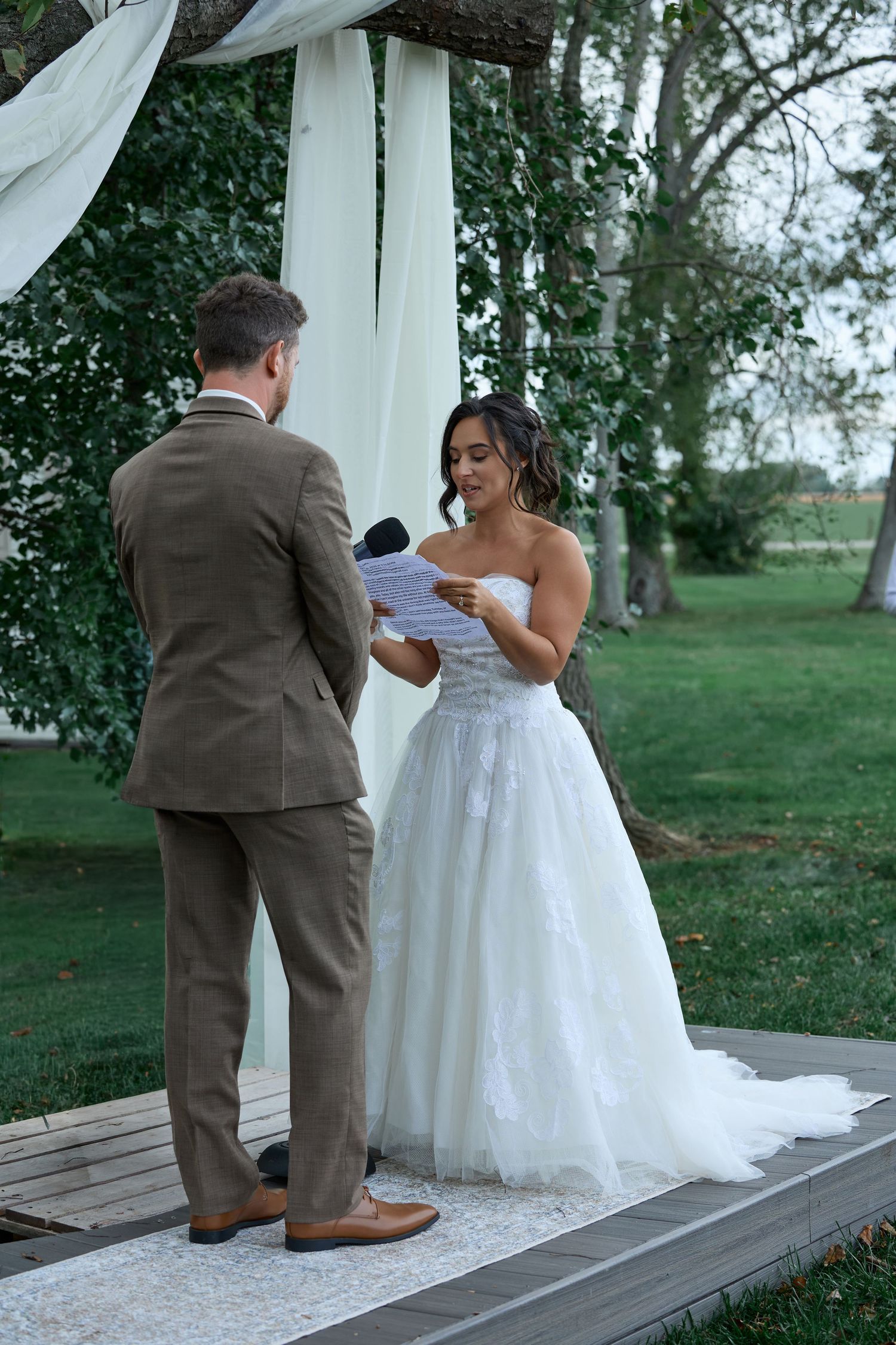 A bride in a strapless white lace gown reads her vows from a paper to the groom under a draped fabric arch outdoors.
