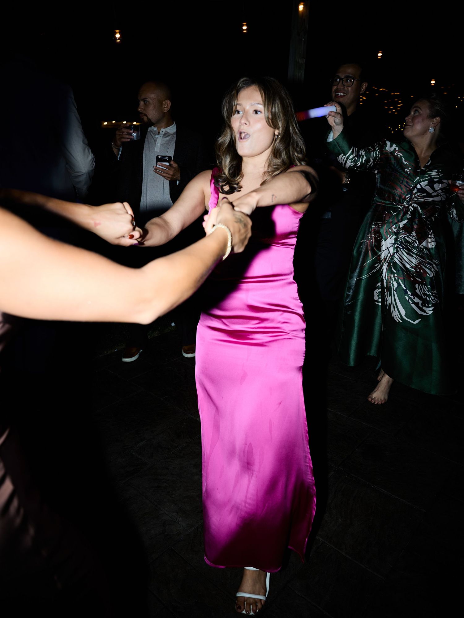 A smiling woman in a bright pink satin dress holds hands with guests while dancing at an evening wedding party.