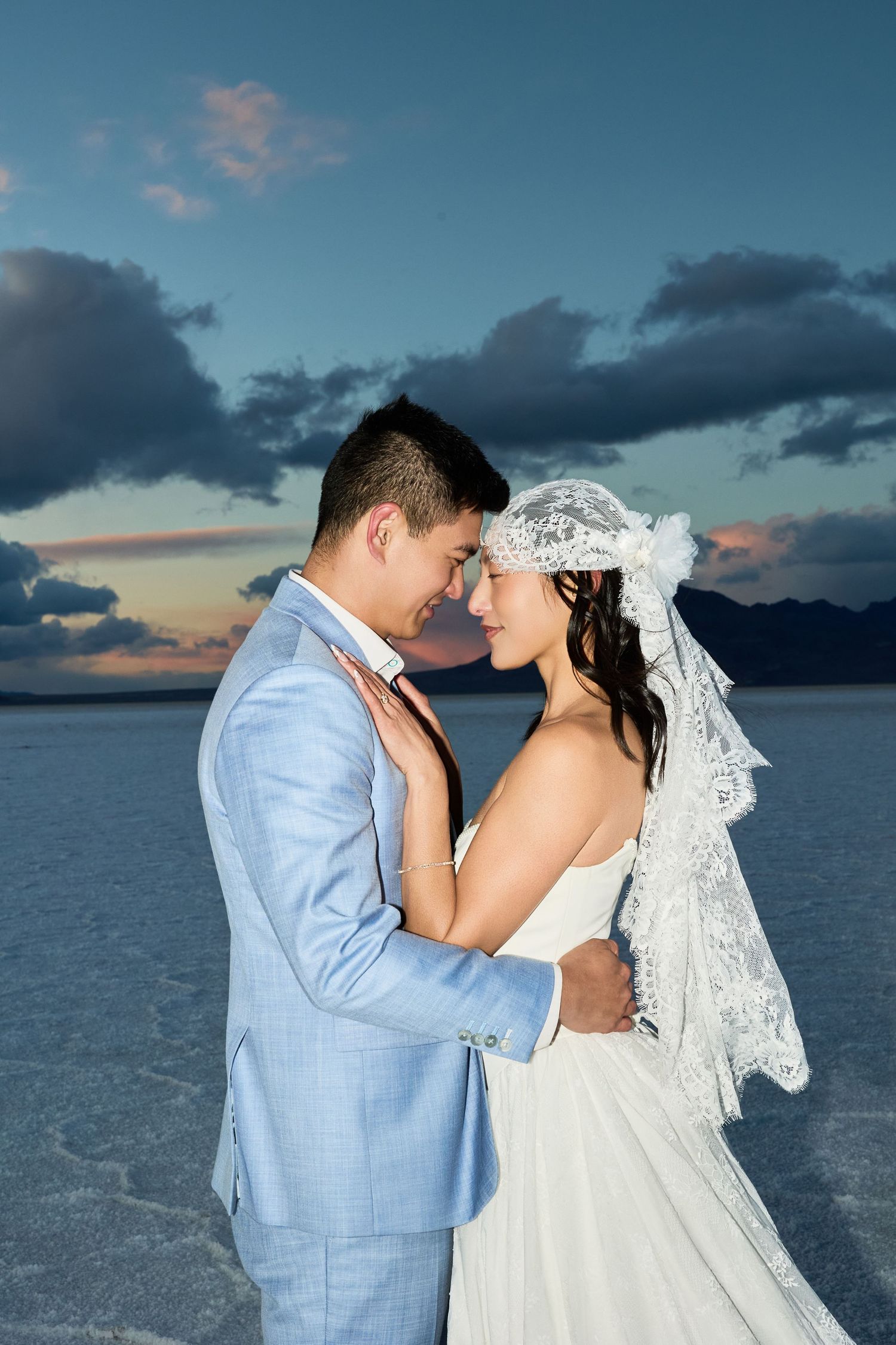 Newlywed couple in a light blue suit and lace veil embrace in the glow of a blue and orange salt flat sunset.