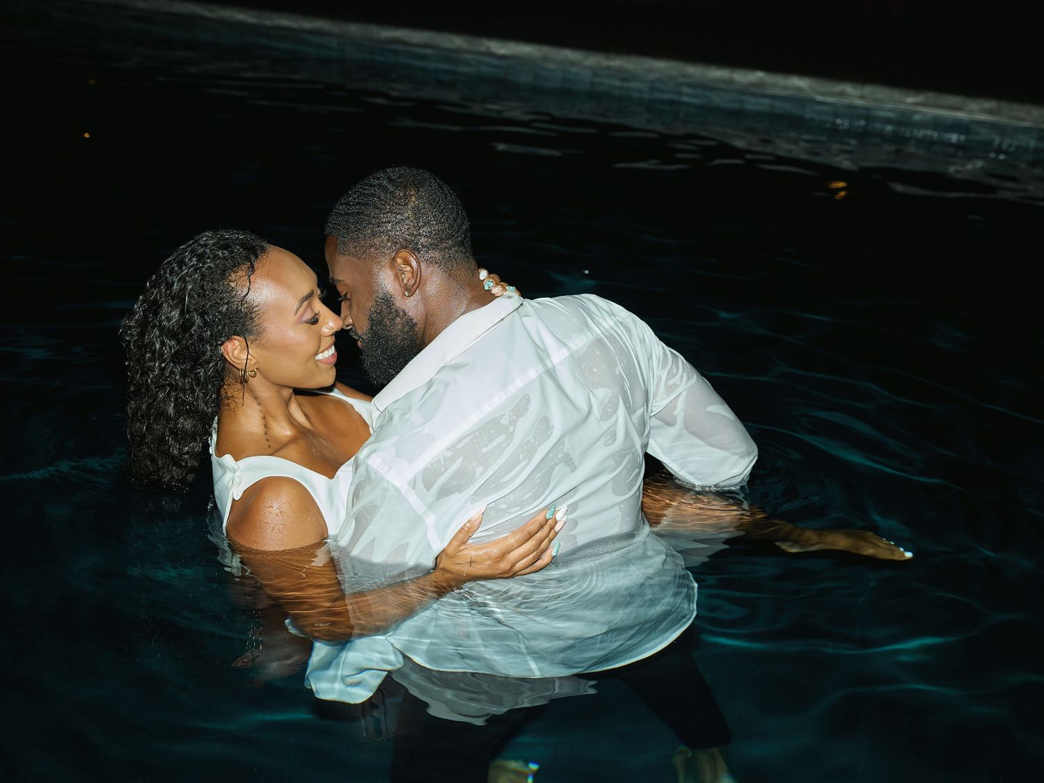 A laughing couple in white outfits embraces in a dark pool at night, enjoying a joyful romantic water photoshoot.