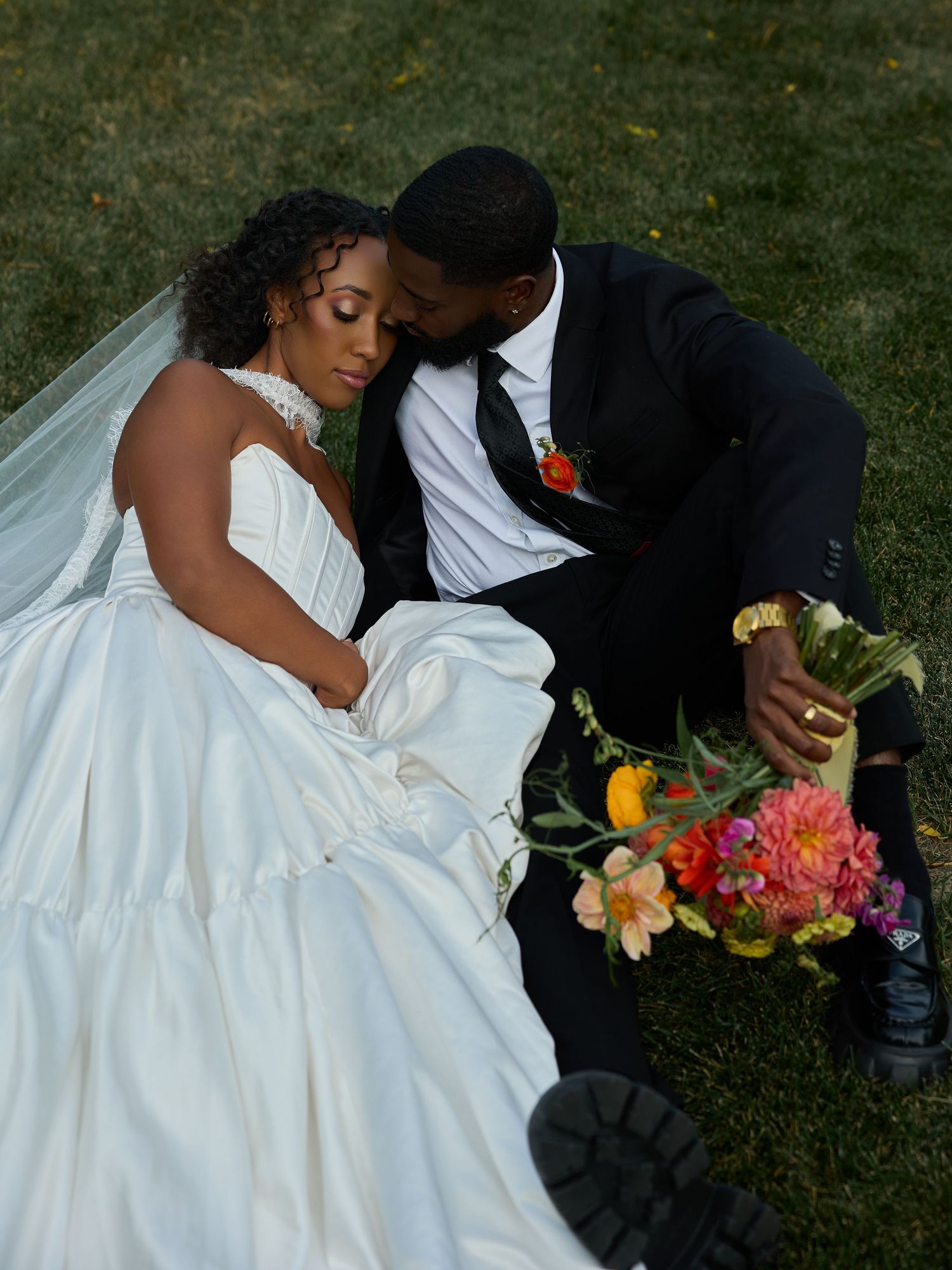Bride in a strapless white gown rests against her groom on the grass, holding a colorful bouquet of pink, orange and yellow flowers.