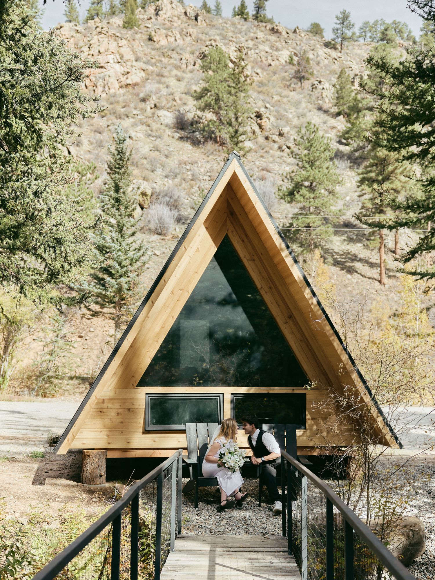 Newlyweds sit on the porch of a modern A-frame cabin with a white bouquet, framed by rocky hills and pine trees.