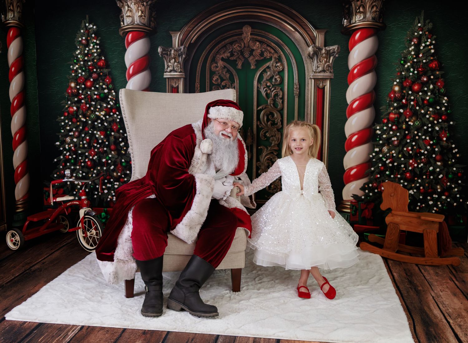 Young girl in white dress and red shoes sits with Santa Claus in festive Christmas setting with decorated trees.