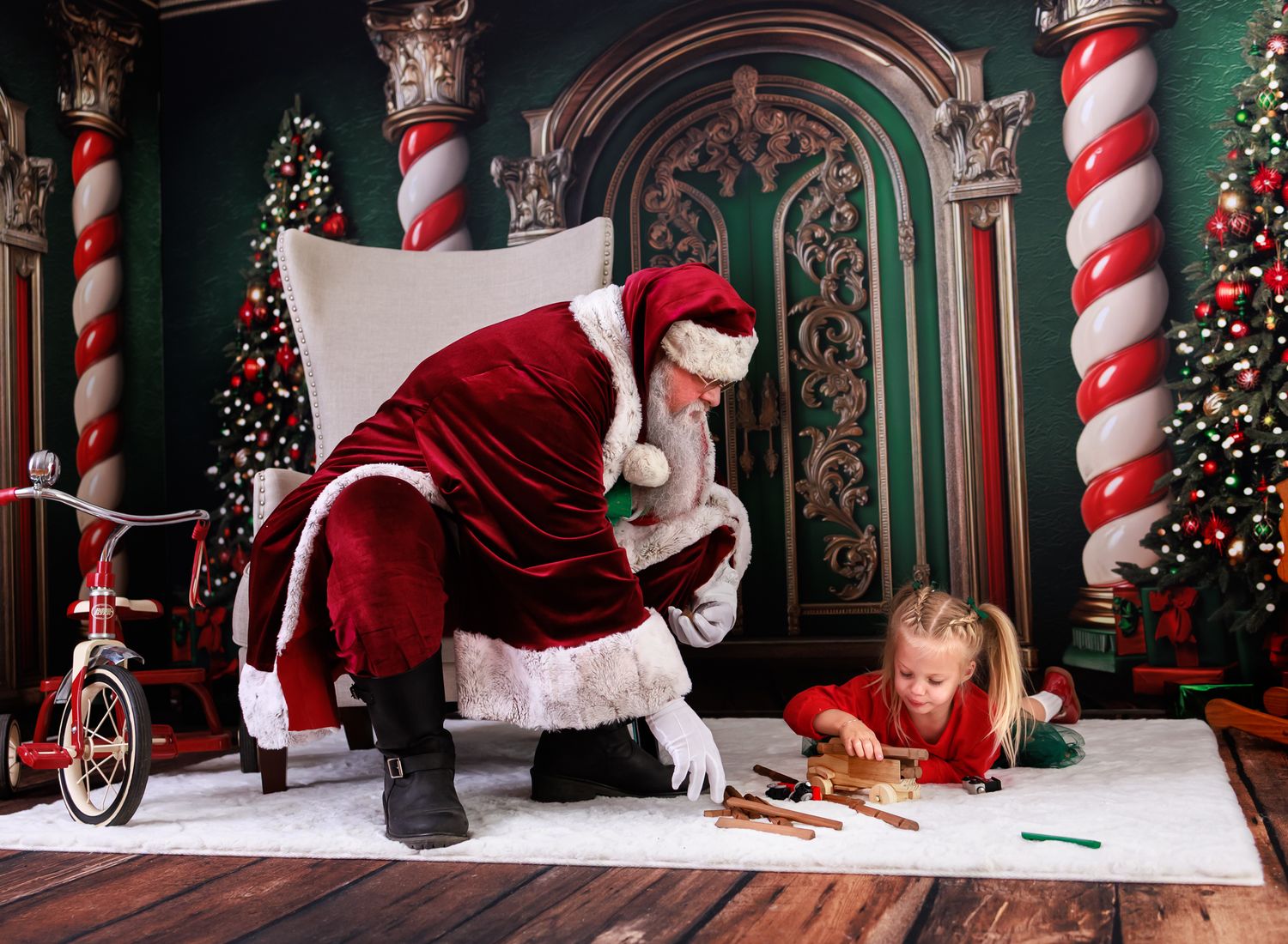 Santa Claus leans over to help a young girl in red write her Christmas wish list on a festive holiday backdrop.