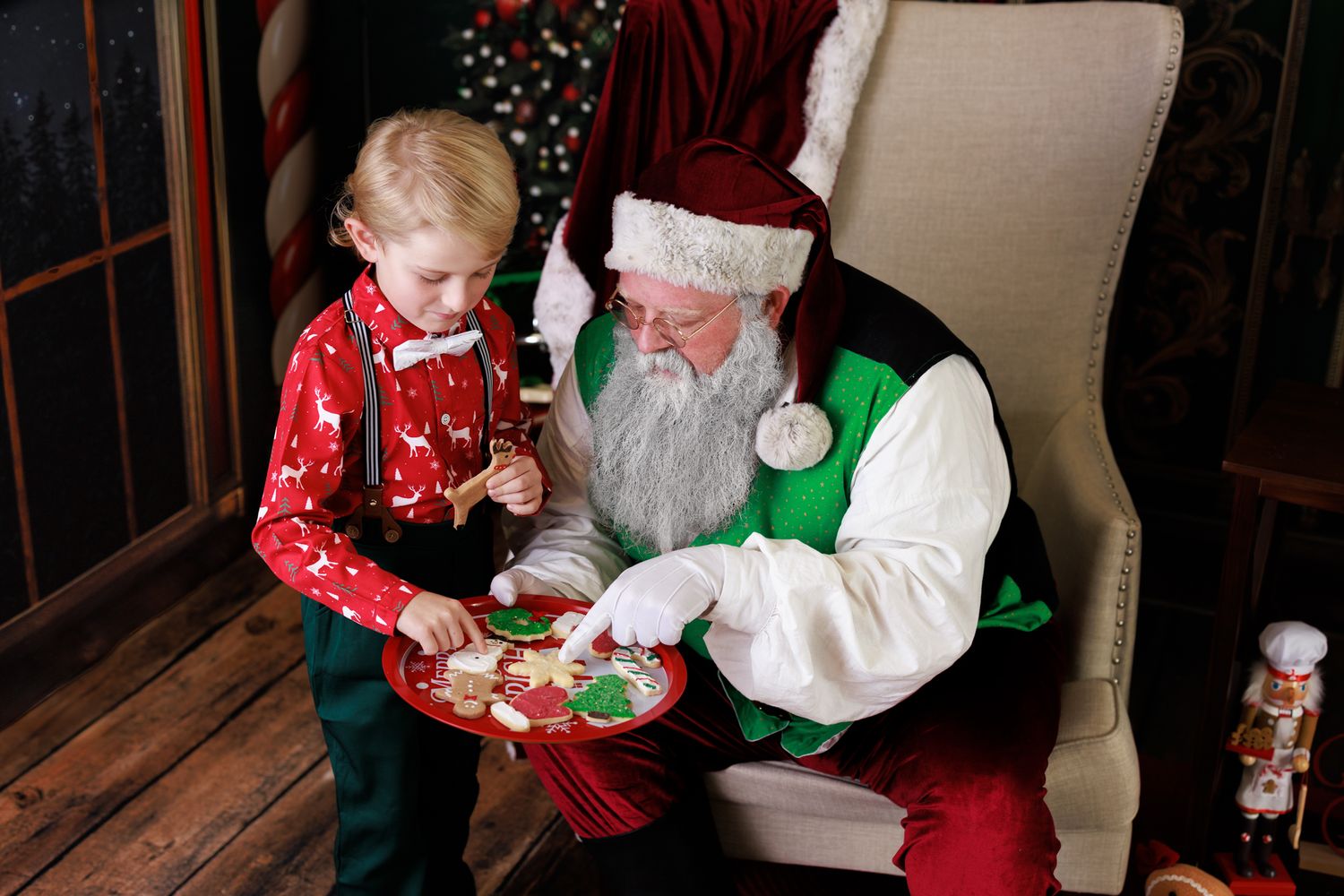 Child in Christmas pajamas sharing cookies with Santa Claus seated in a festive chair during a holiday photo session.