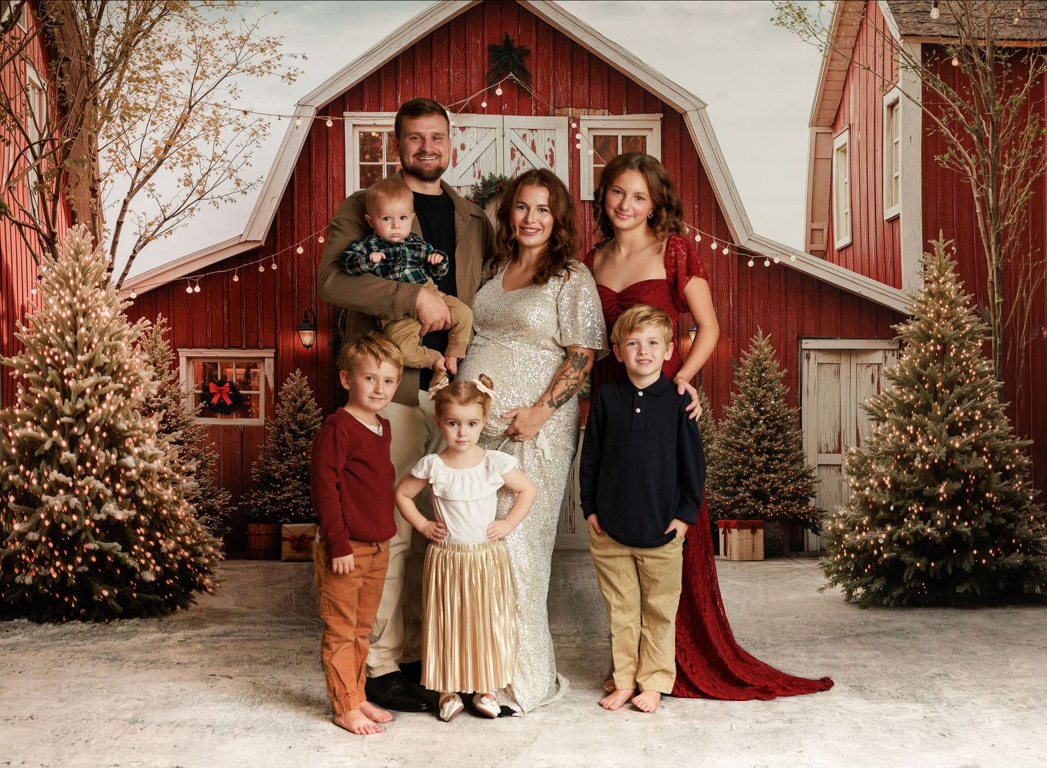 Family of six posing for a Christmas portrait in front of a red barn with holiday trees and festive decor.