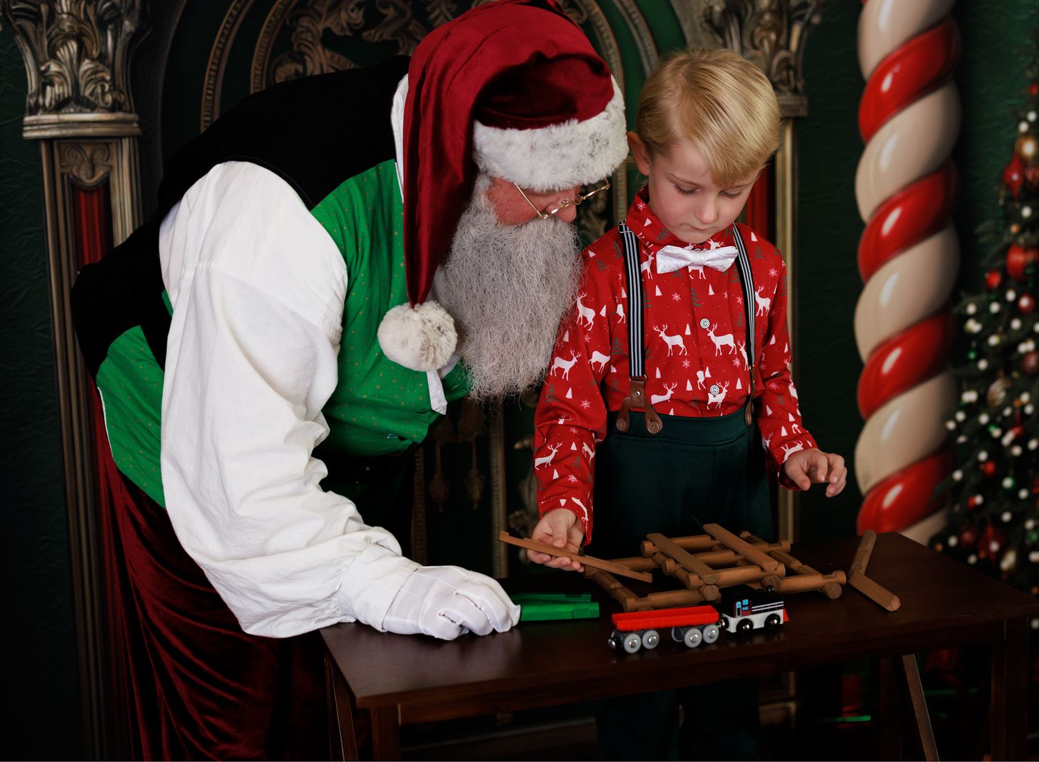 Santa Claus showing a young boy a wooden toy train set during a festive Christmas photo session.