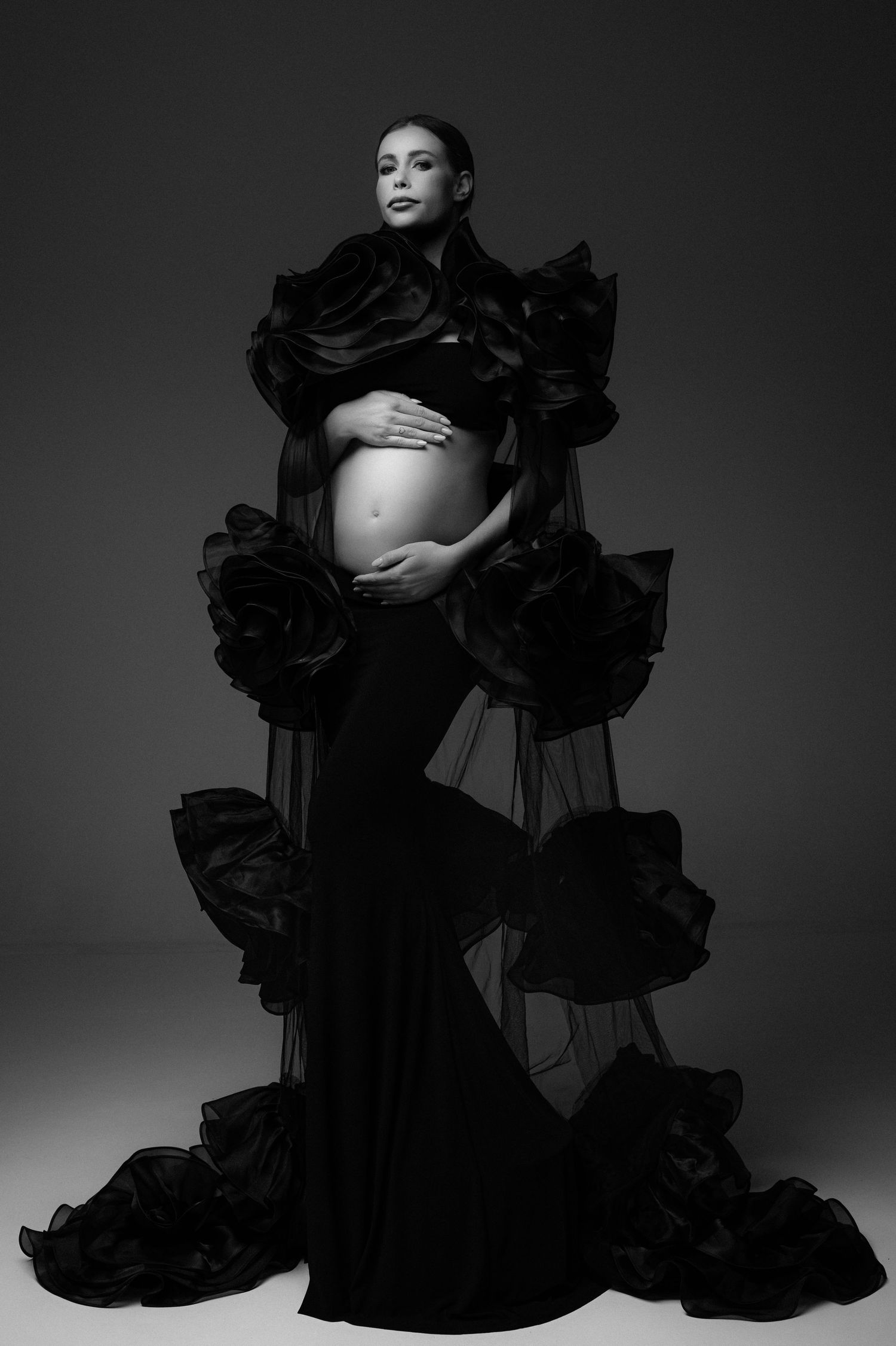 Pregnant woman in elegant black ruffled gown with floral details, cradling baby bump in dramatic B&W maternity portrait.