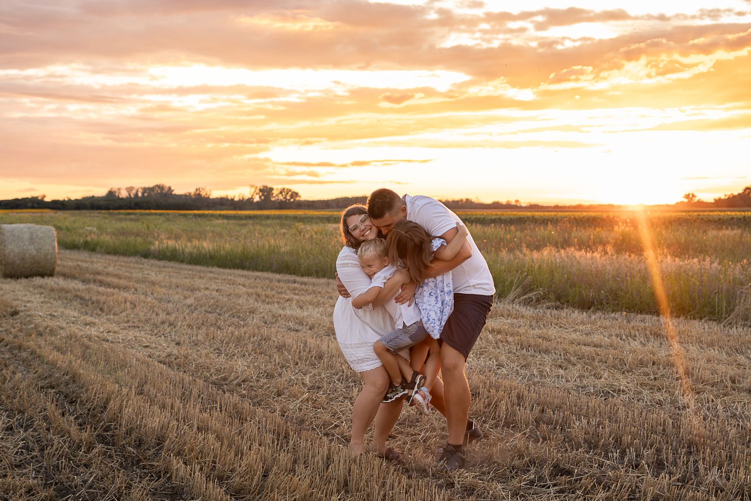 Family hugging in a golden wheat field at sunset with hay bales in the background.