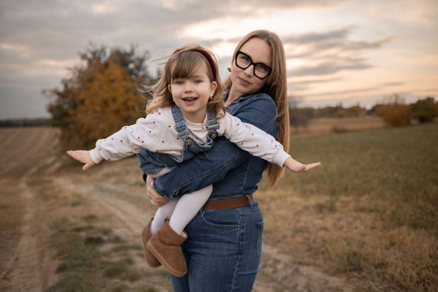 A mother holds her smiling young daughter outdoors in a field at sunset, both wearing denim outfits.