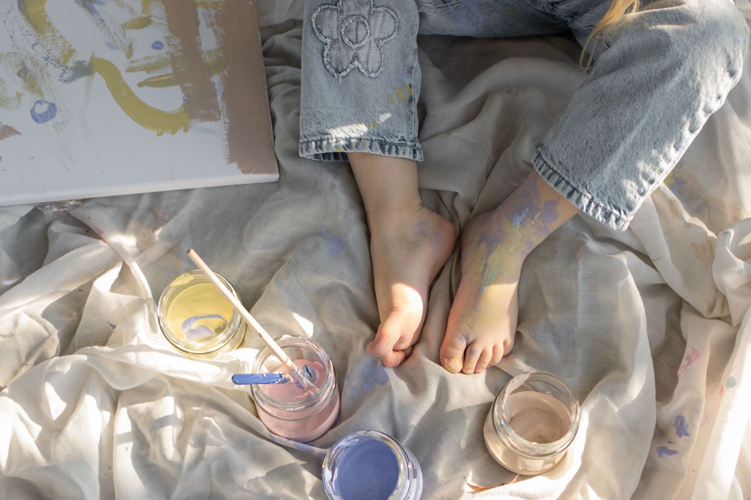 Child sitting on white fabric with painted feet, surrounded by colorful paint jars and a paintbrush nearby.