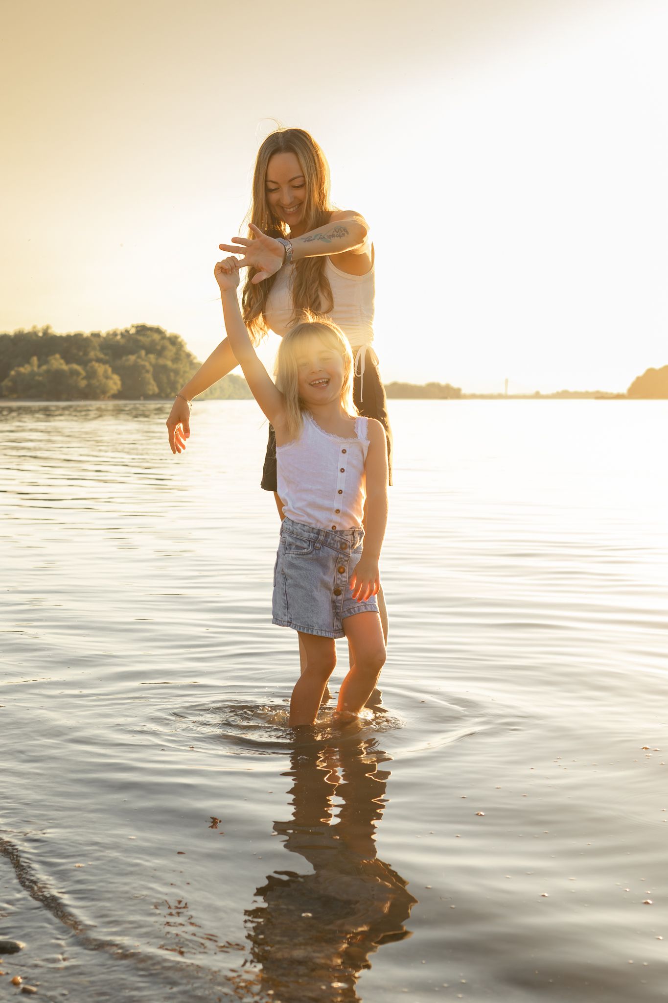 Mother and daughter playing in shallow water at sunset, smiling and holding hands on a warm summer evening.