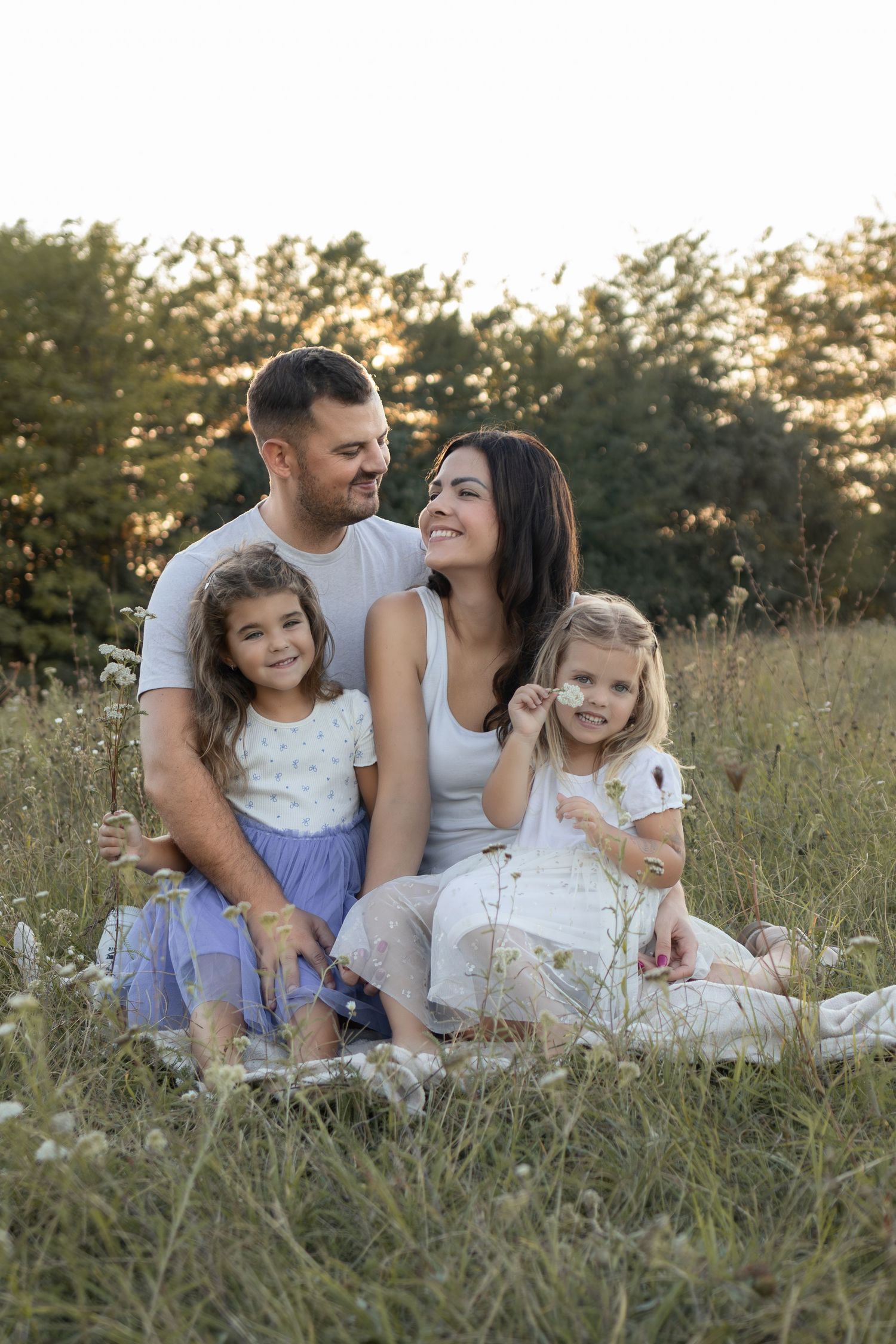 Happy family of four sitting in a sunny meadow with wildflowers, parents and two young daughters smiling outdoors.