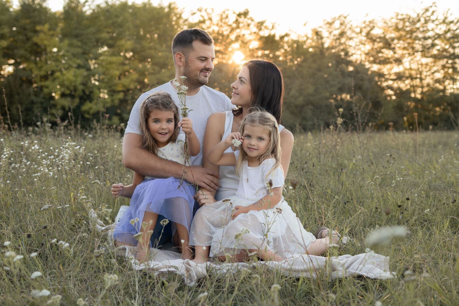 Family of four sitting in a sunny meadow with wildflowers, parents and two daughters smiling warmly at golden hour.