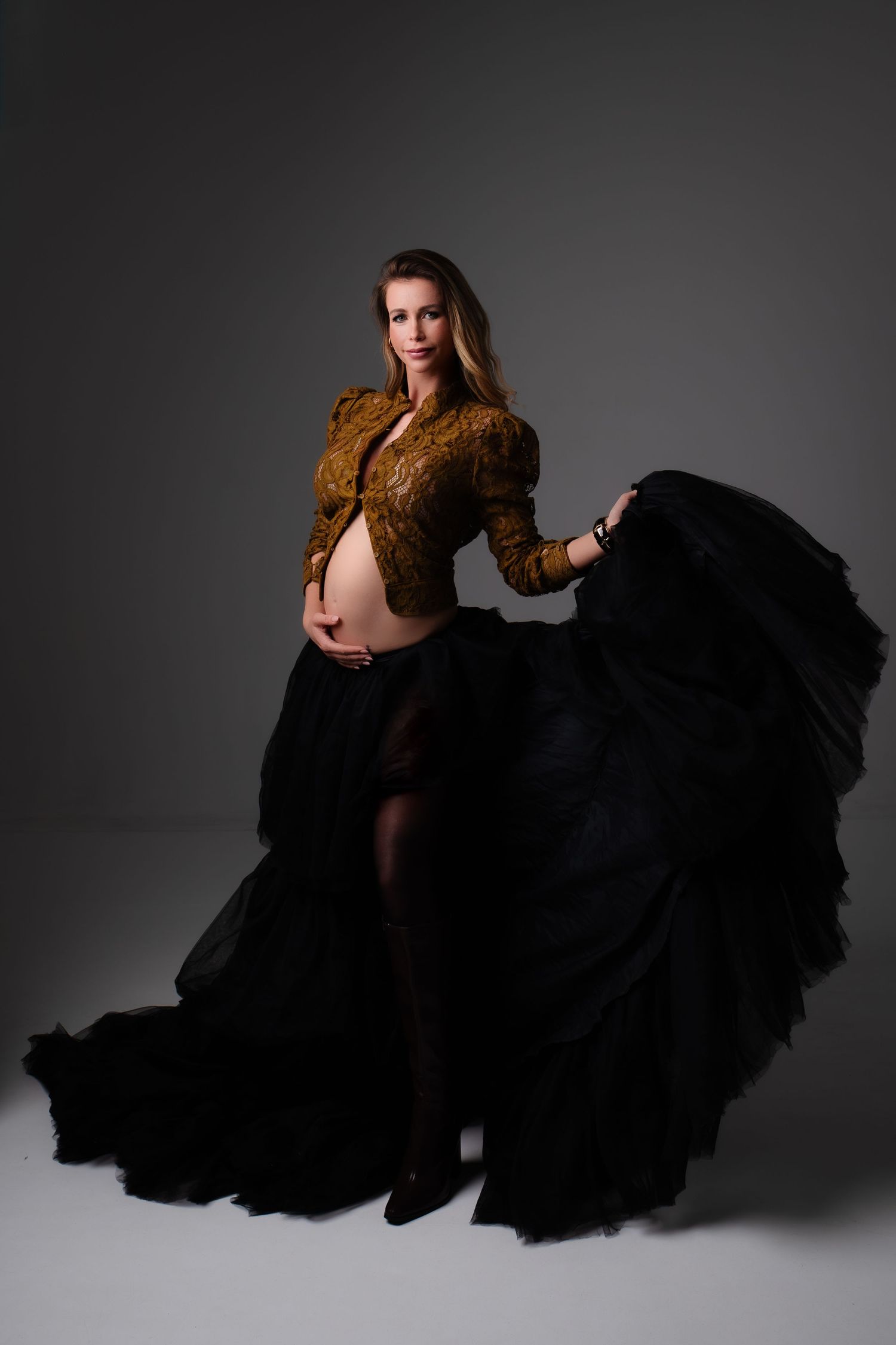 Pregnant woman in gold crop top and flowing black tulle skirt posing elegantly in studio maternity photo shoot.