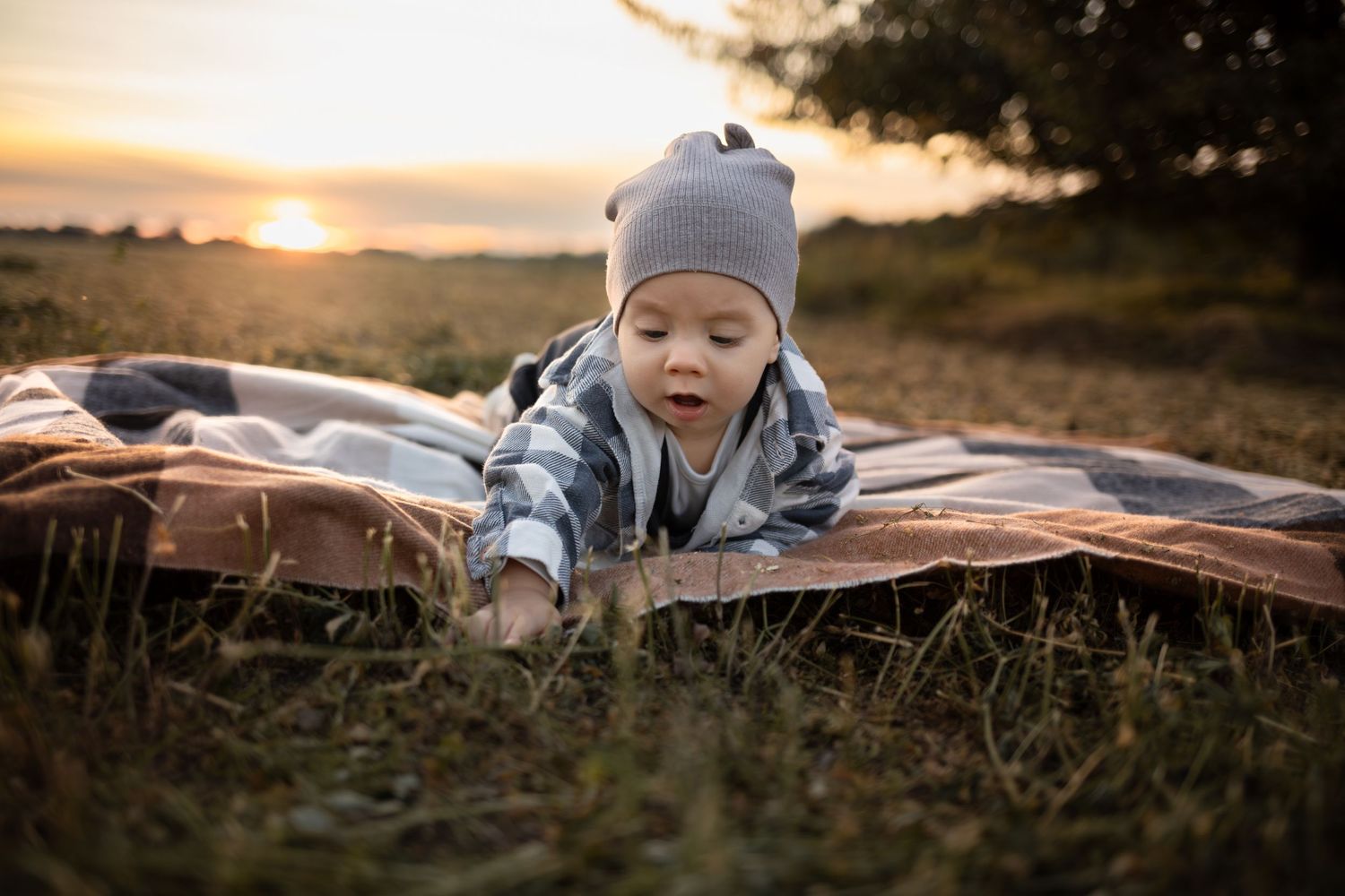 A curious baby in a gray beanie lies on a blanket in a field during a golden sunset outdoors.