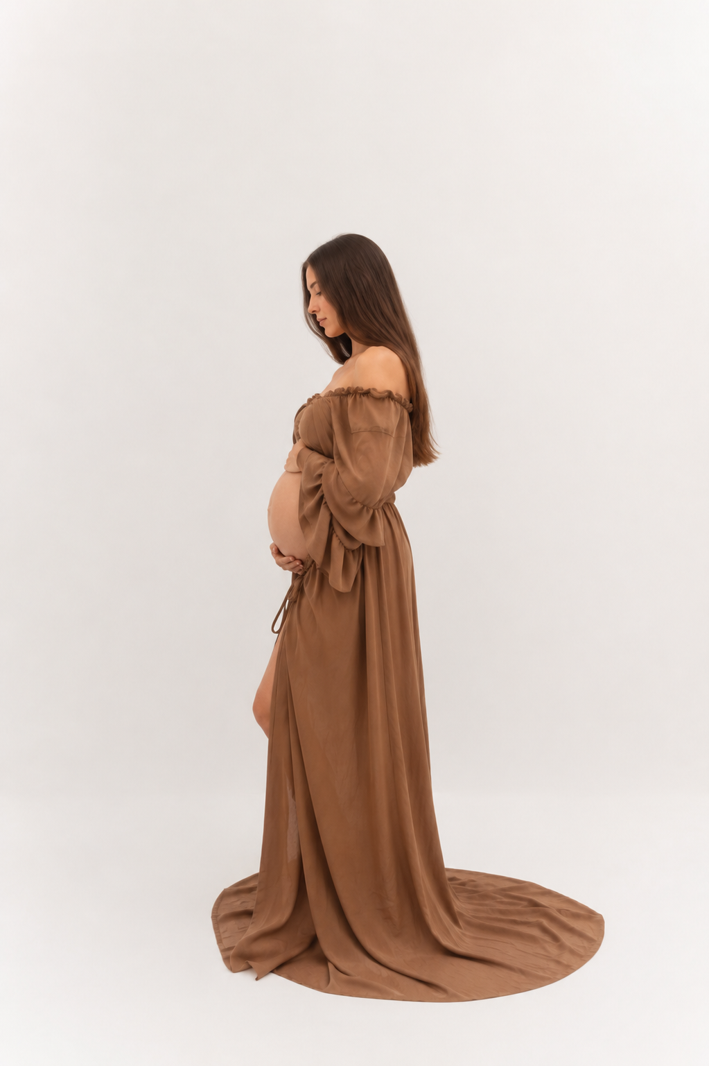 Pregnant woman in elegant brown off-shoulder maternity gown with flowing train poses sideways in studio.