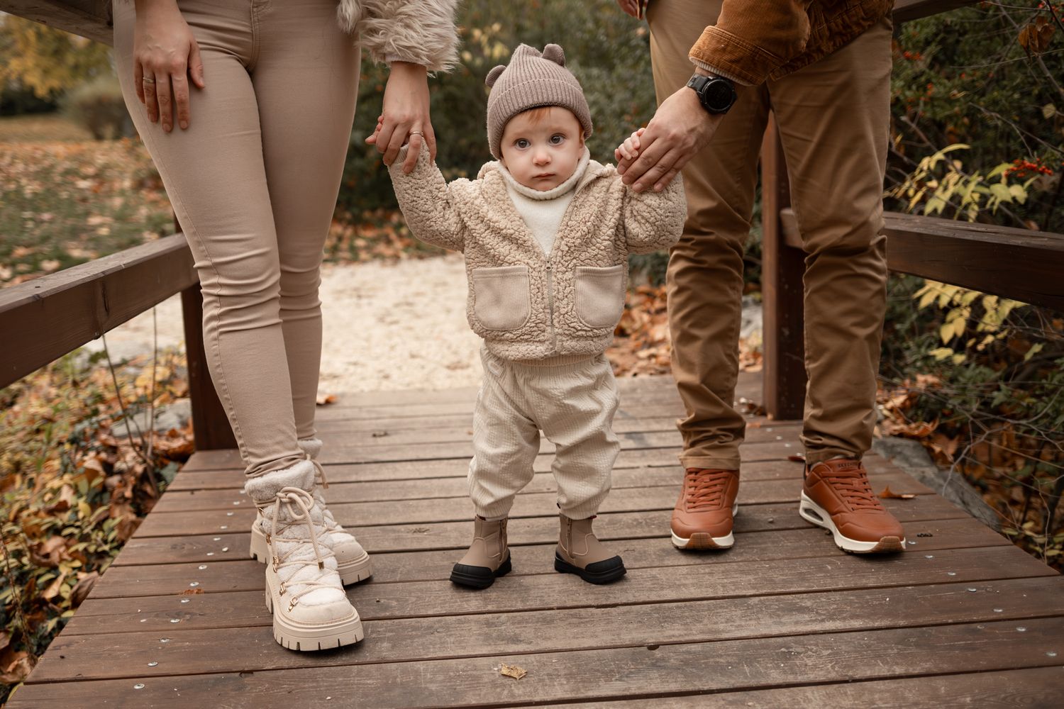Baby in beige knit outfit stands on wooden bridge held by parents in autumn park.