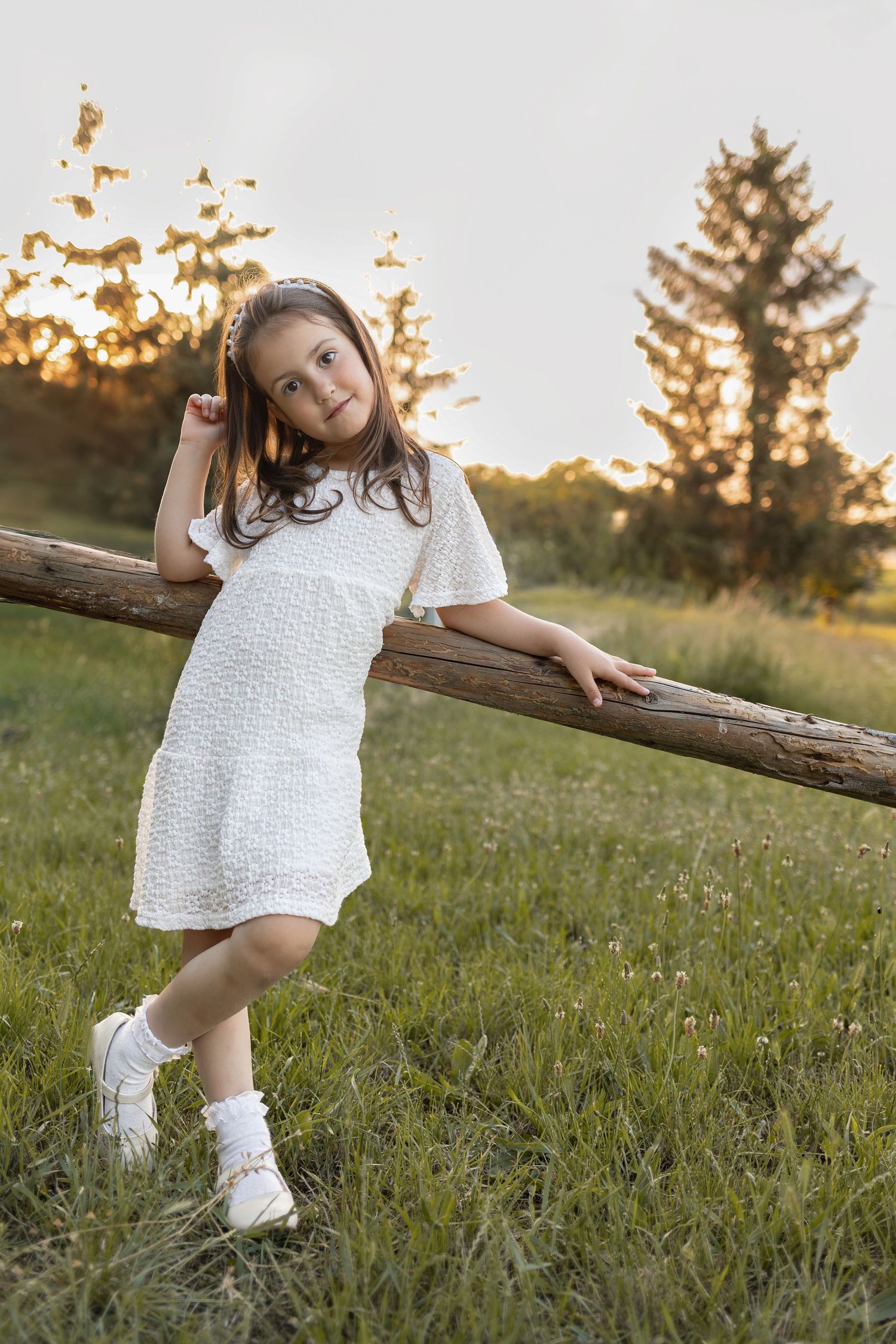 Young girl in white dress leaning on wooden fence rail in sunny green meadow at golden hour sunset.