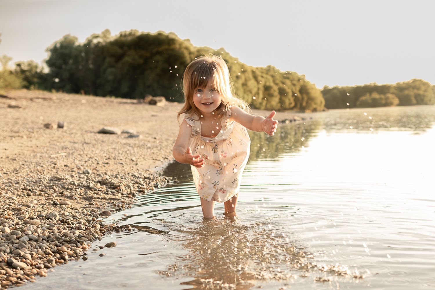 A joyful toddler girl splashing in shallow river water on a sunny pebble beach surrounded by green trees.