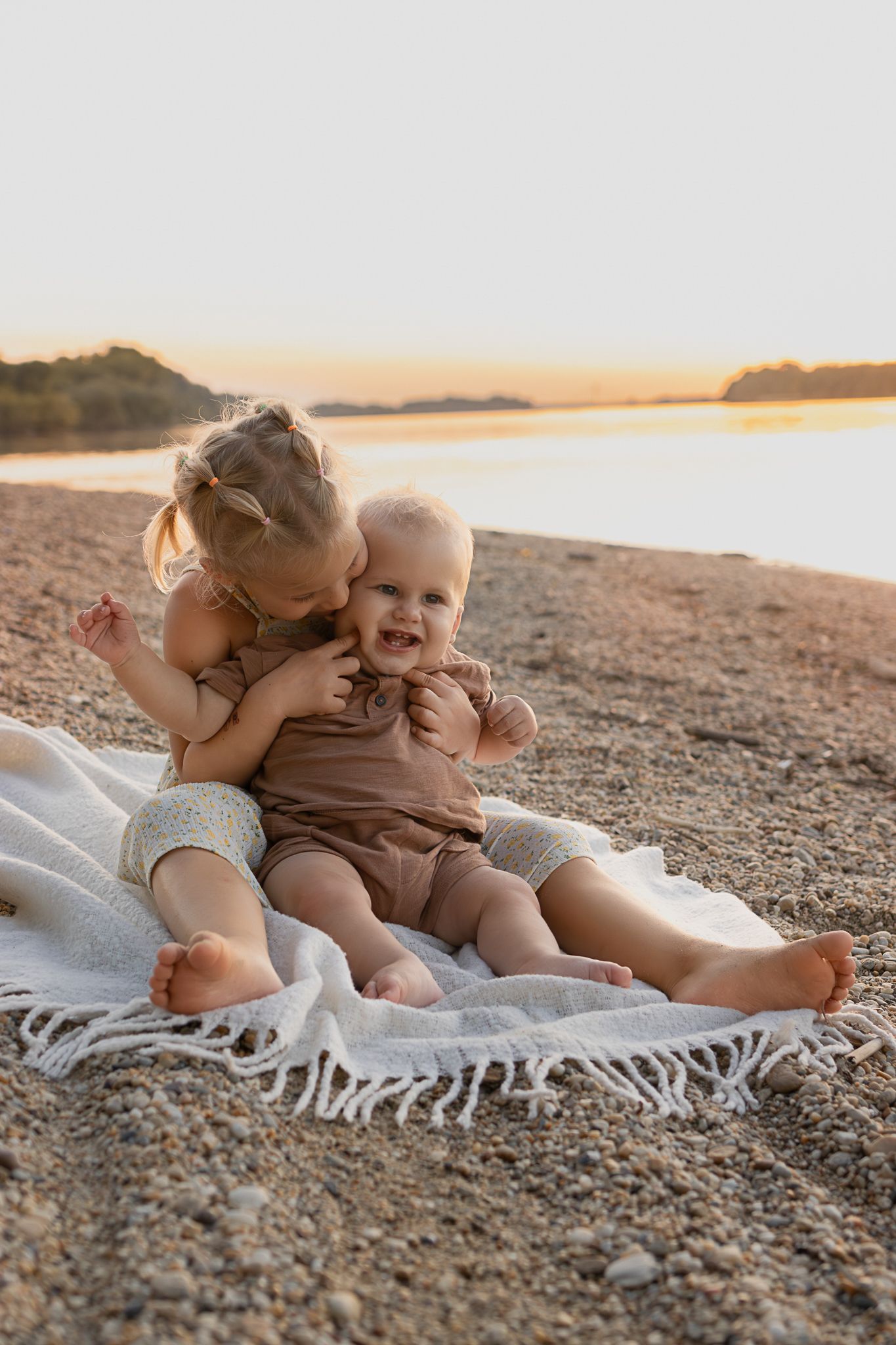 Two young siblings sitting on a blanket at a sandy beach during golden hour sunset, older girl kissing baby.