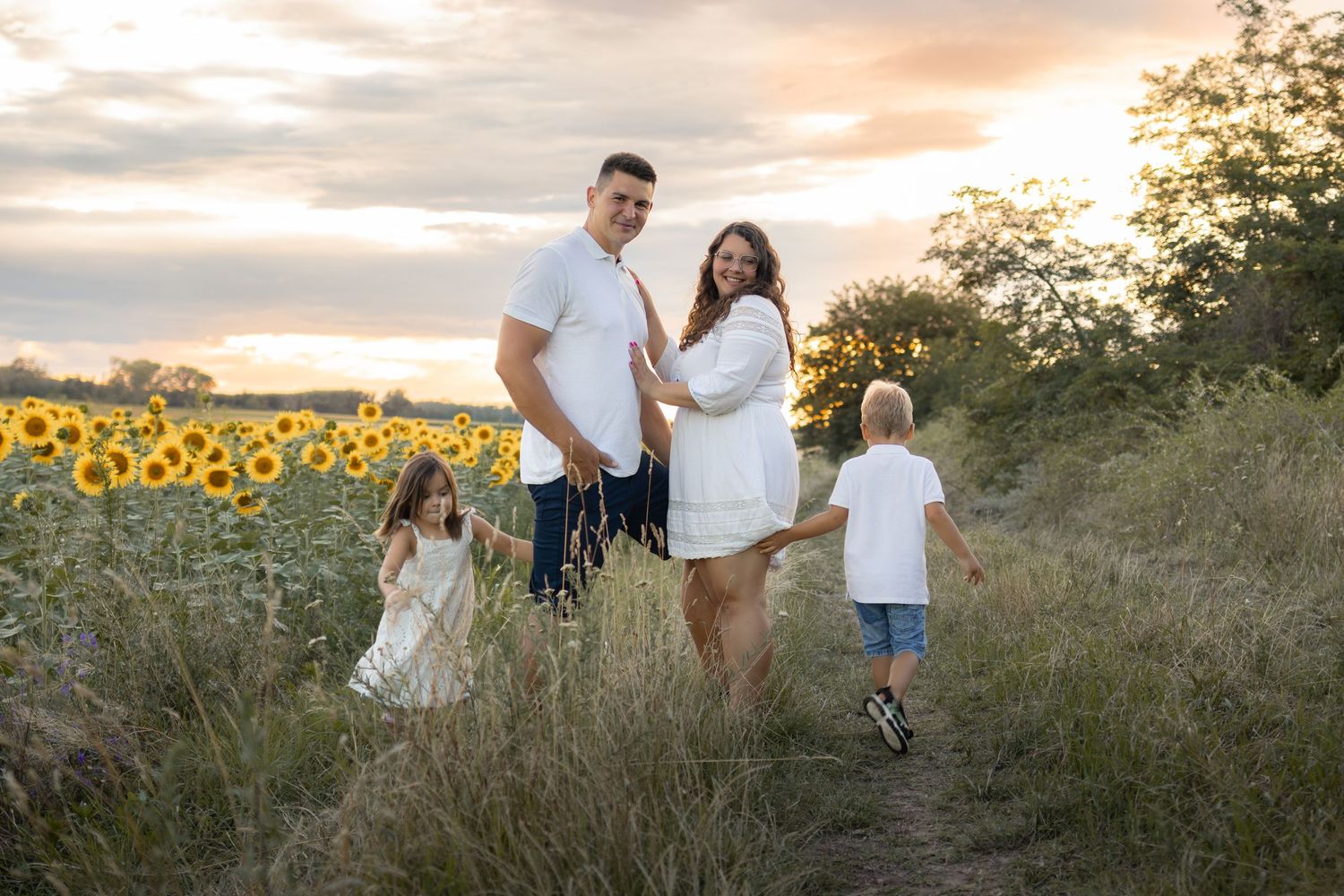 Family of four poses in a sunflower field at sunset, parents holding hands with young children on a dirt path.