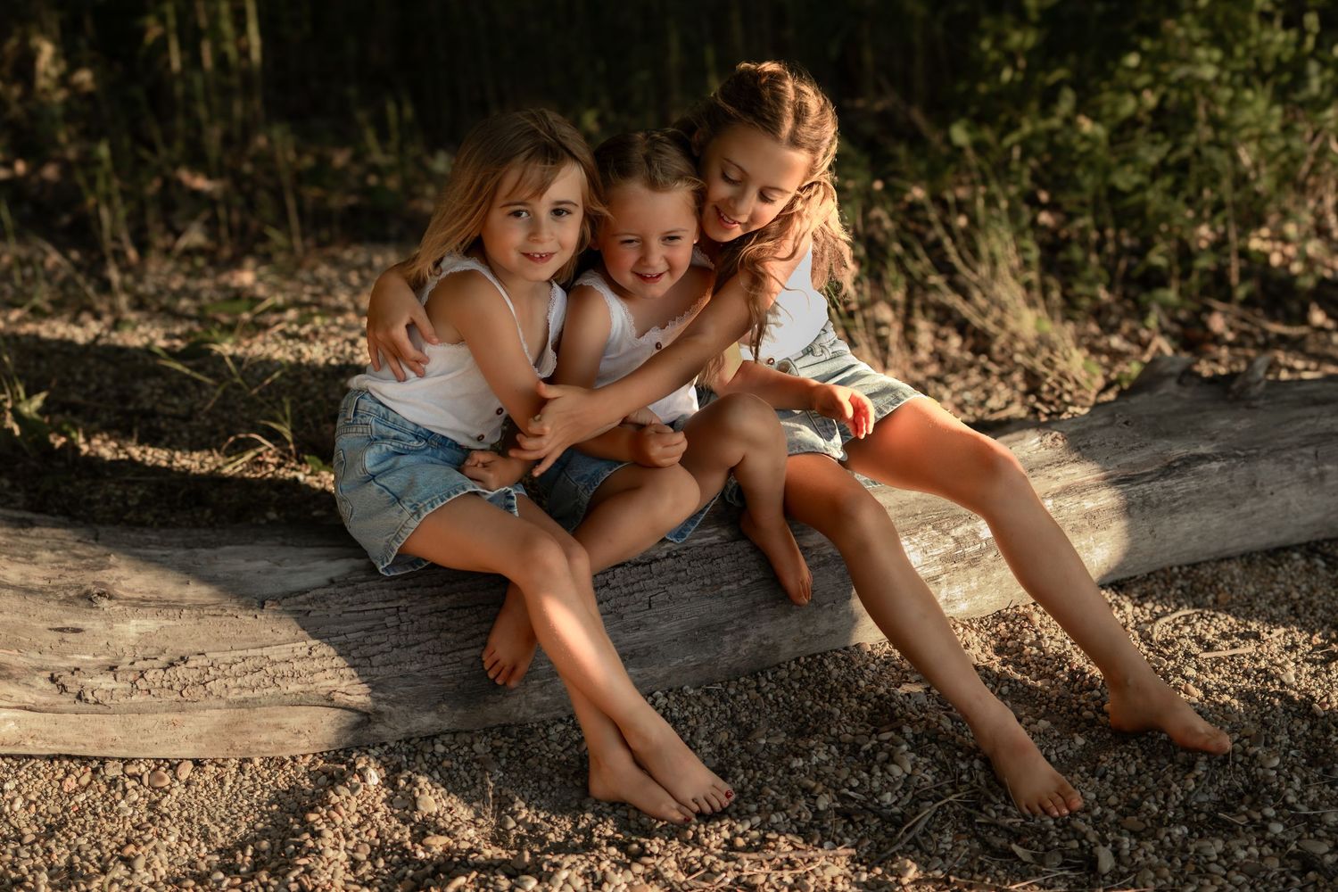 Three young girls sitting together on a log outdoors, smiling and hugging in warm golden sunlight.