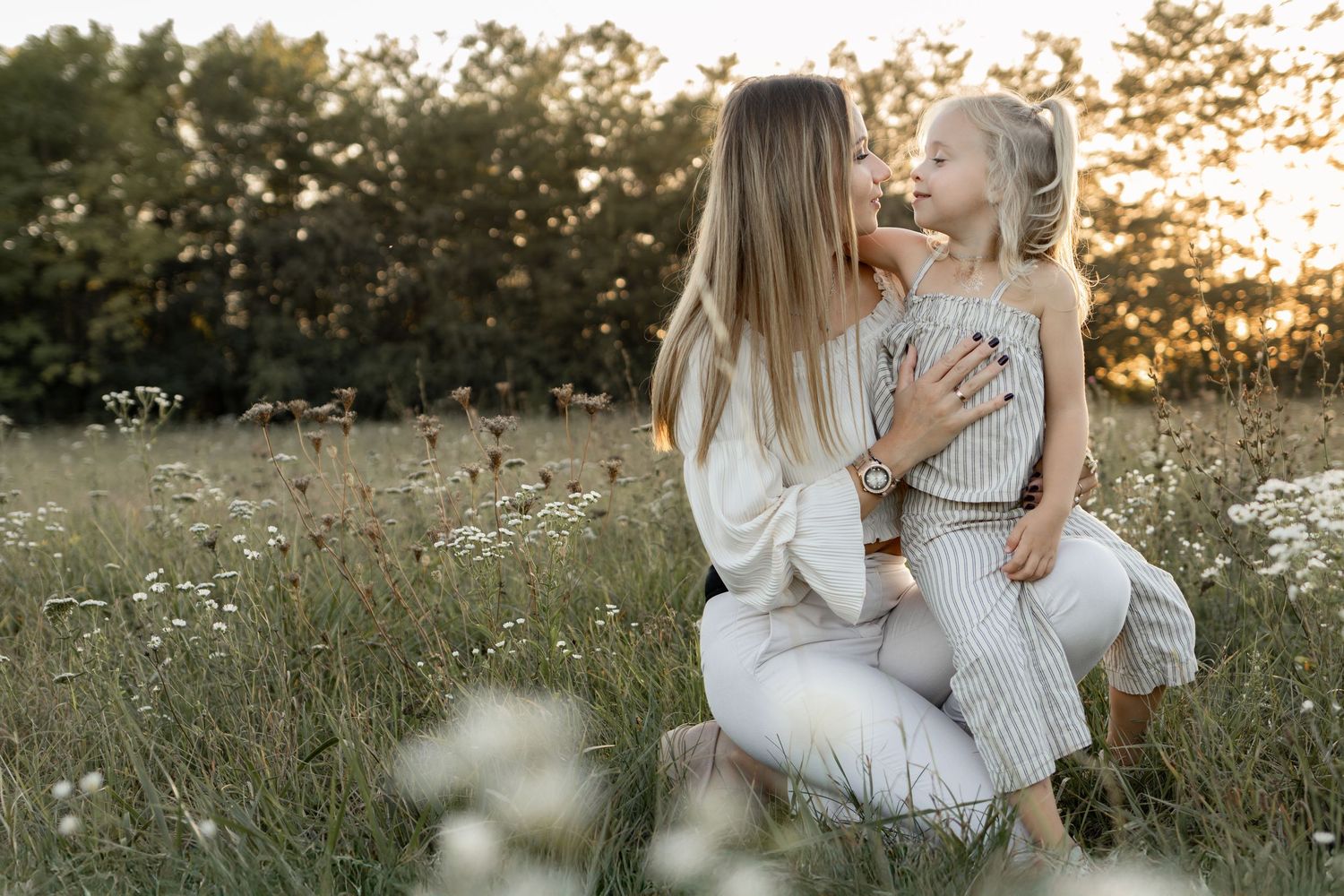 A mother crouches in a wildflower field at sunset, sharing a tender moment with her smiling young daughter.
