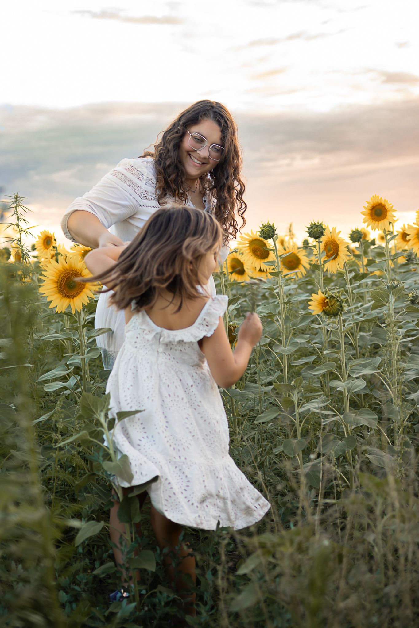 Mother and daughter dancing joyfully in a sunflower field at sunset, both wearing white dresses.