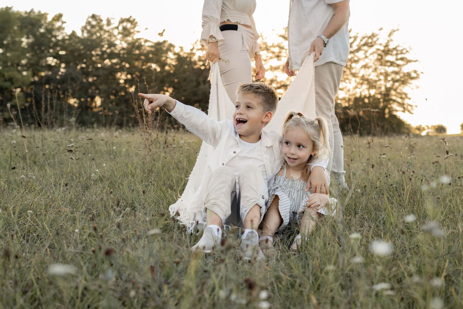 Two happy children sit in a sunny field while their parents stand behind them during a family photo session.