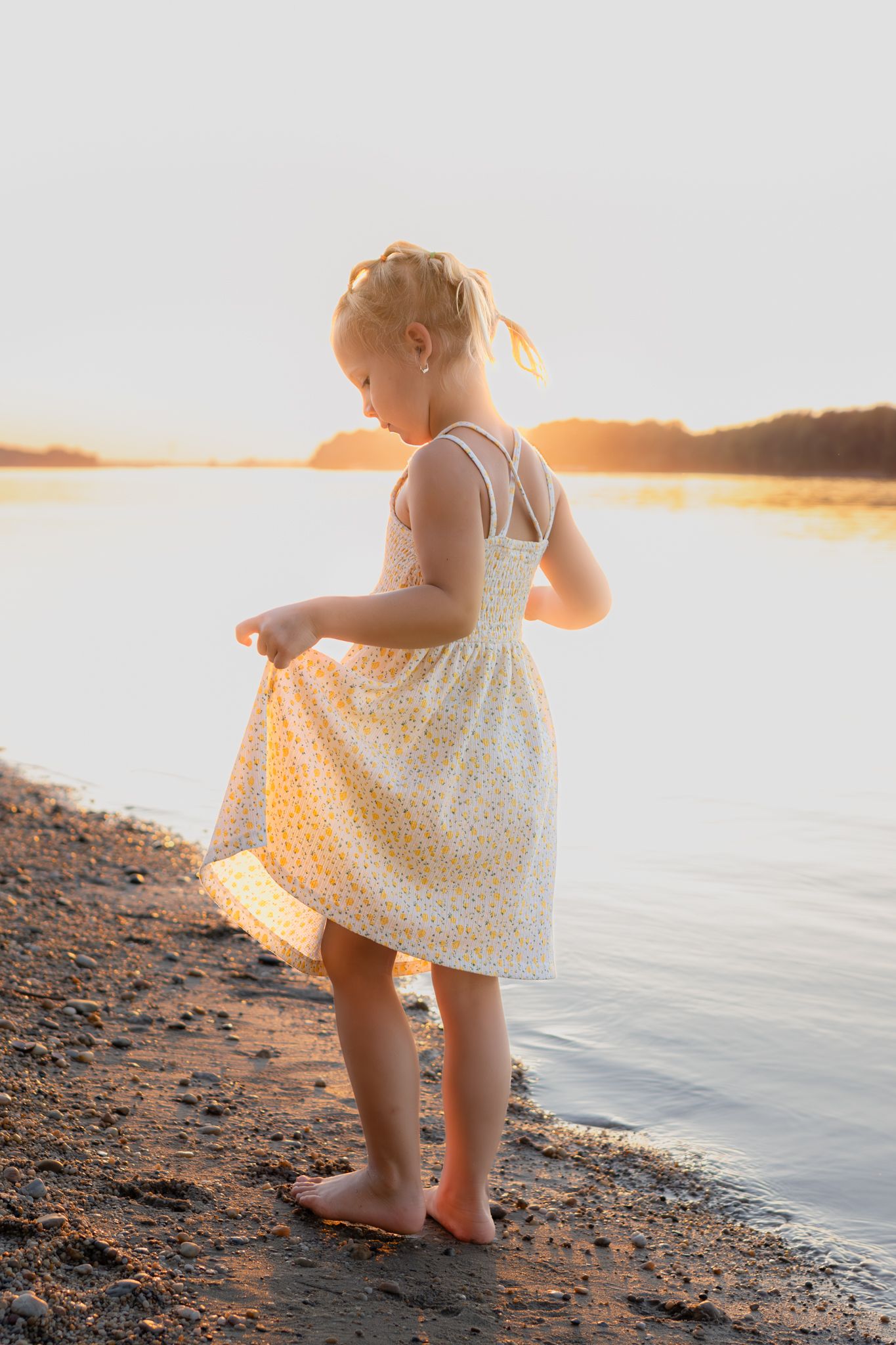 A barefoot little girl in a floral sundress stands by a calm lake shore during a golden sunset.