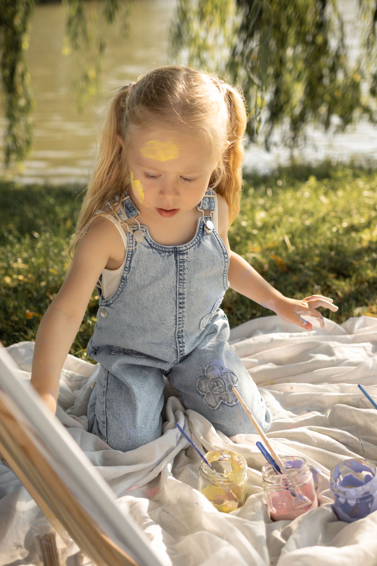 Child painting outdoors on blanket under willow tree branches.