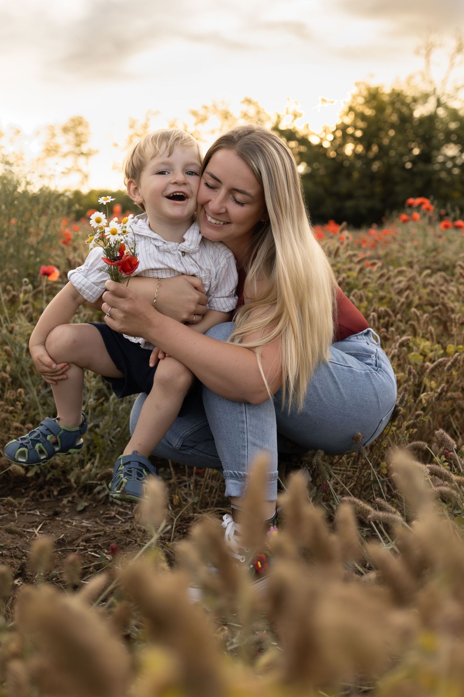 A smiling mother hugs her laughing toddler son in a sunny wildflower field, both holding a small bouquet.