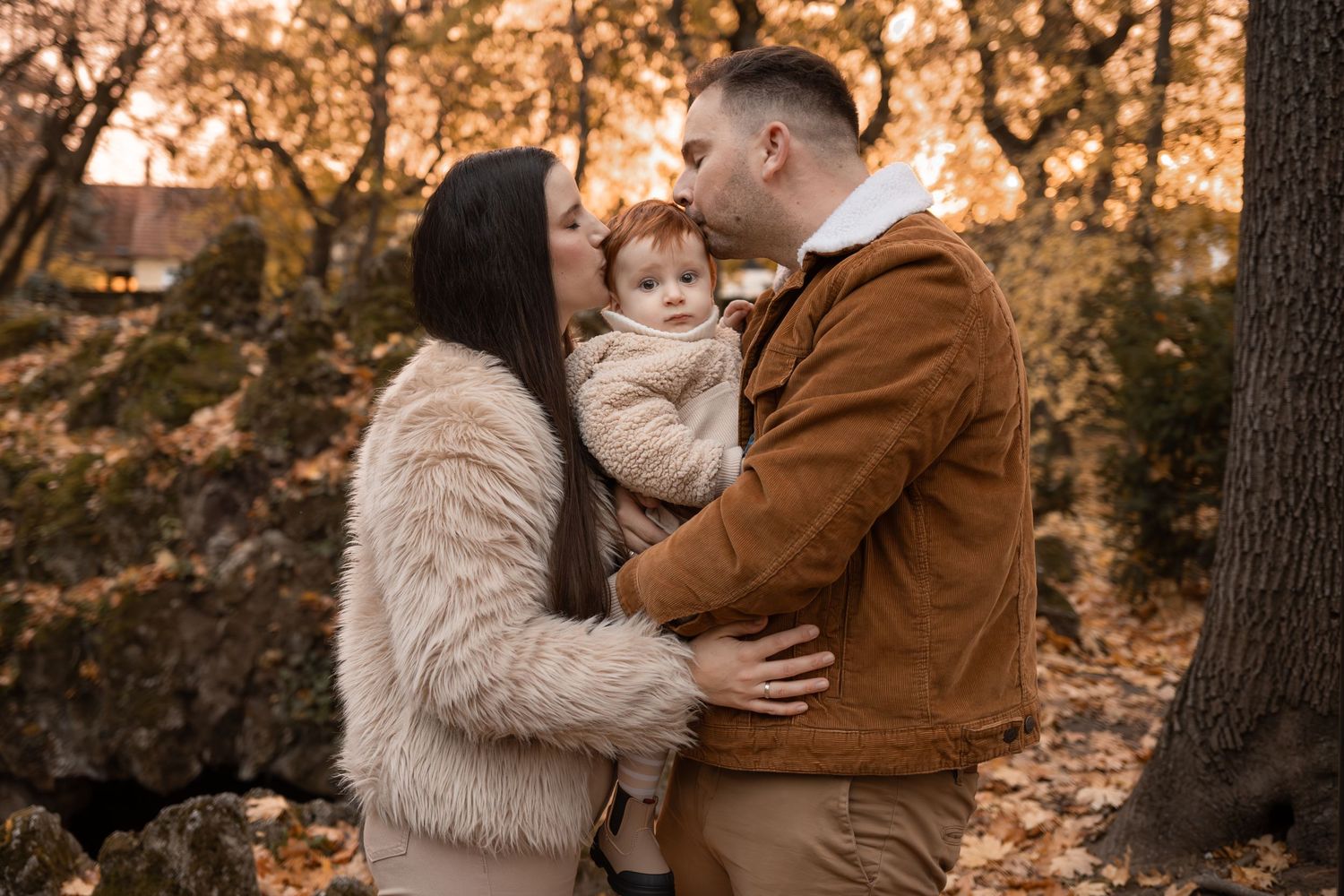 A couple shares a kiss over their baby during an autumn family portrait session in a forest setting.