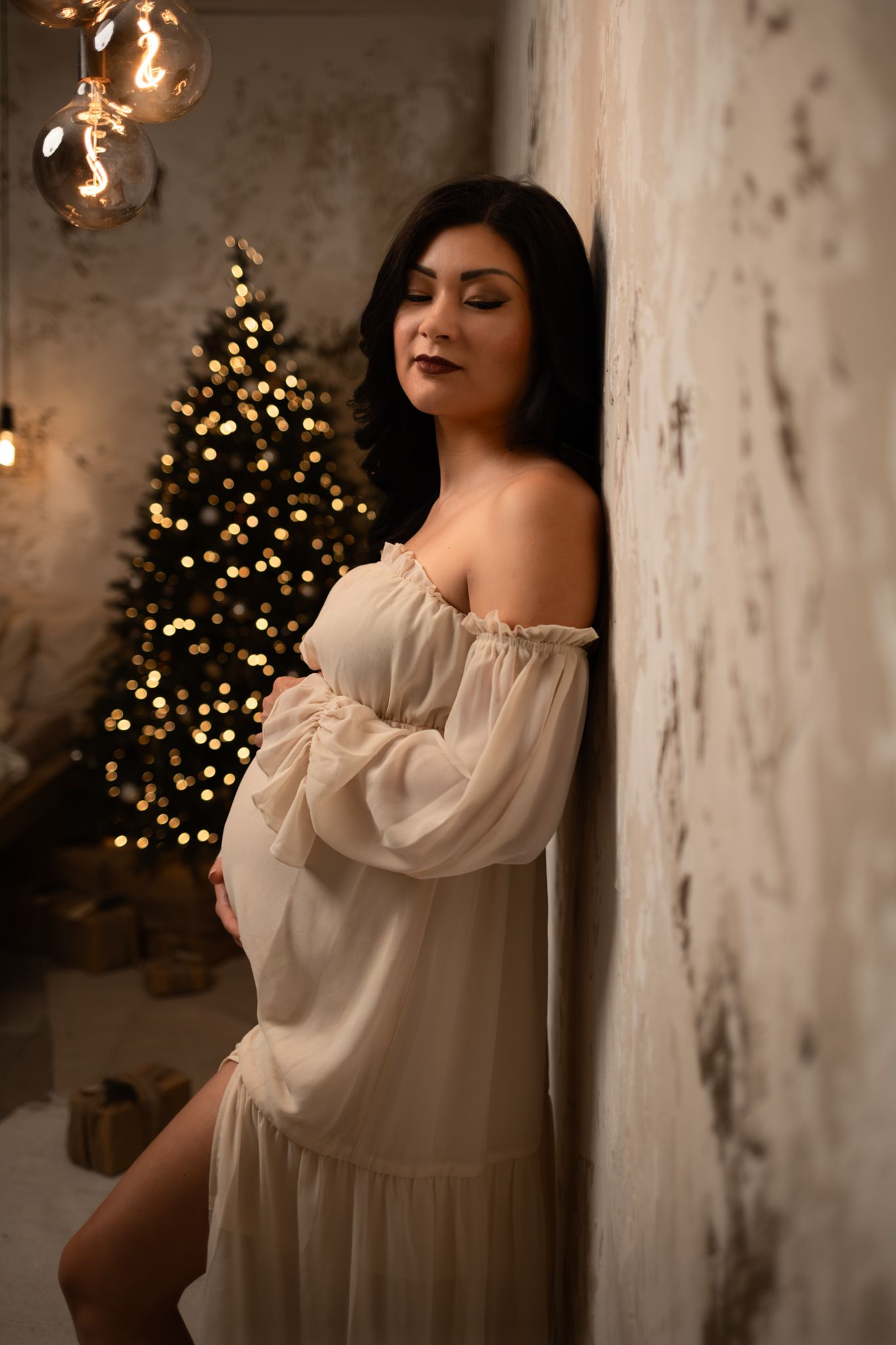 A woman in a flowing white dress poses gracefully against a wall with a Christmas tree and twinkling lights in the background.