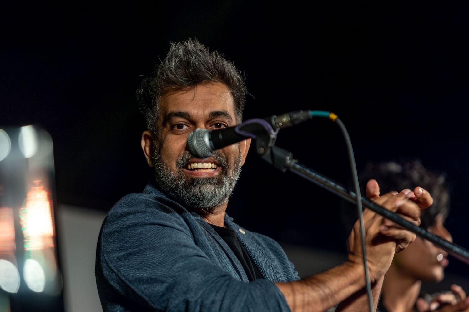 Performer in blue shirt singing passionately into microphone on darkened stage with dramatic lighting.