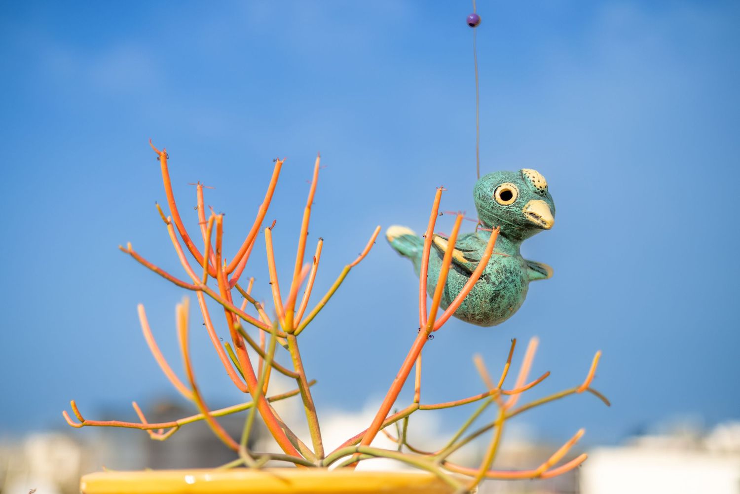 Small green toy figure sitting on orange branch against blue sky.