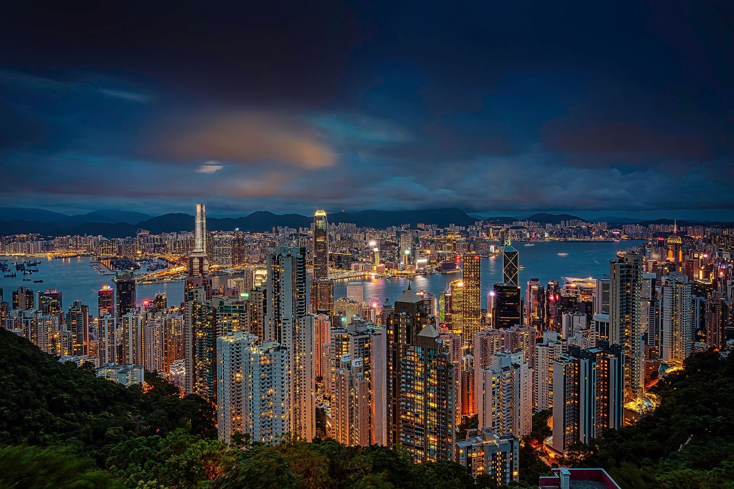 Hong Kong harbor cityscape at dusk with lit skyscrapers against dramatic blue sky.