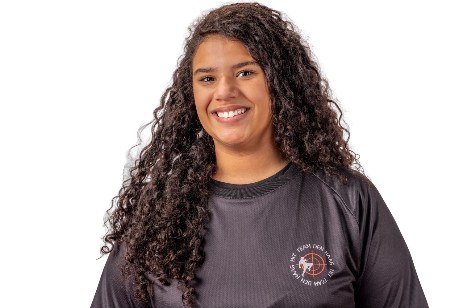 A person with long curly dark hair wearing a grey t-shirt smiles warmly at the camera.