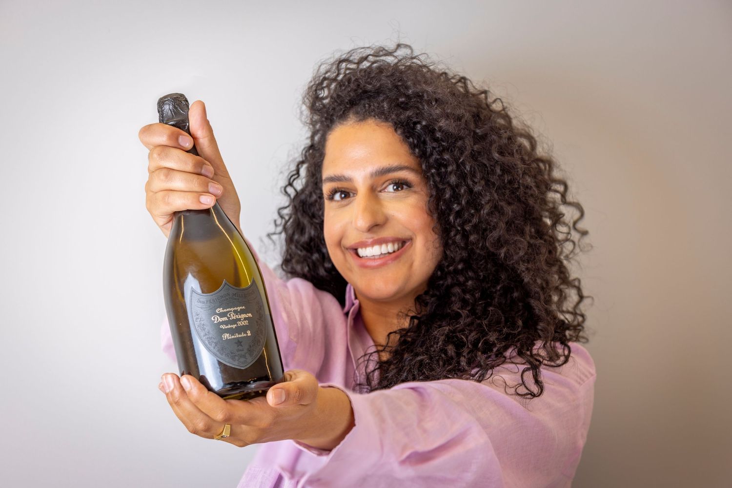 A person in a pink shirt holds up a bottle of champagne with curly dark hair and a warm expression.