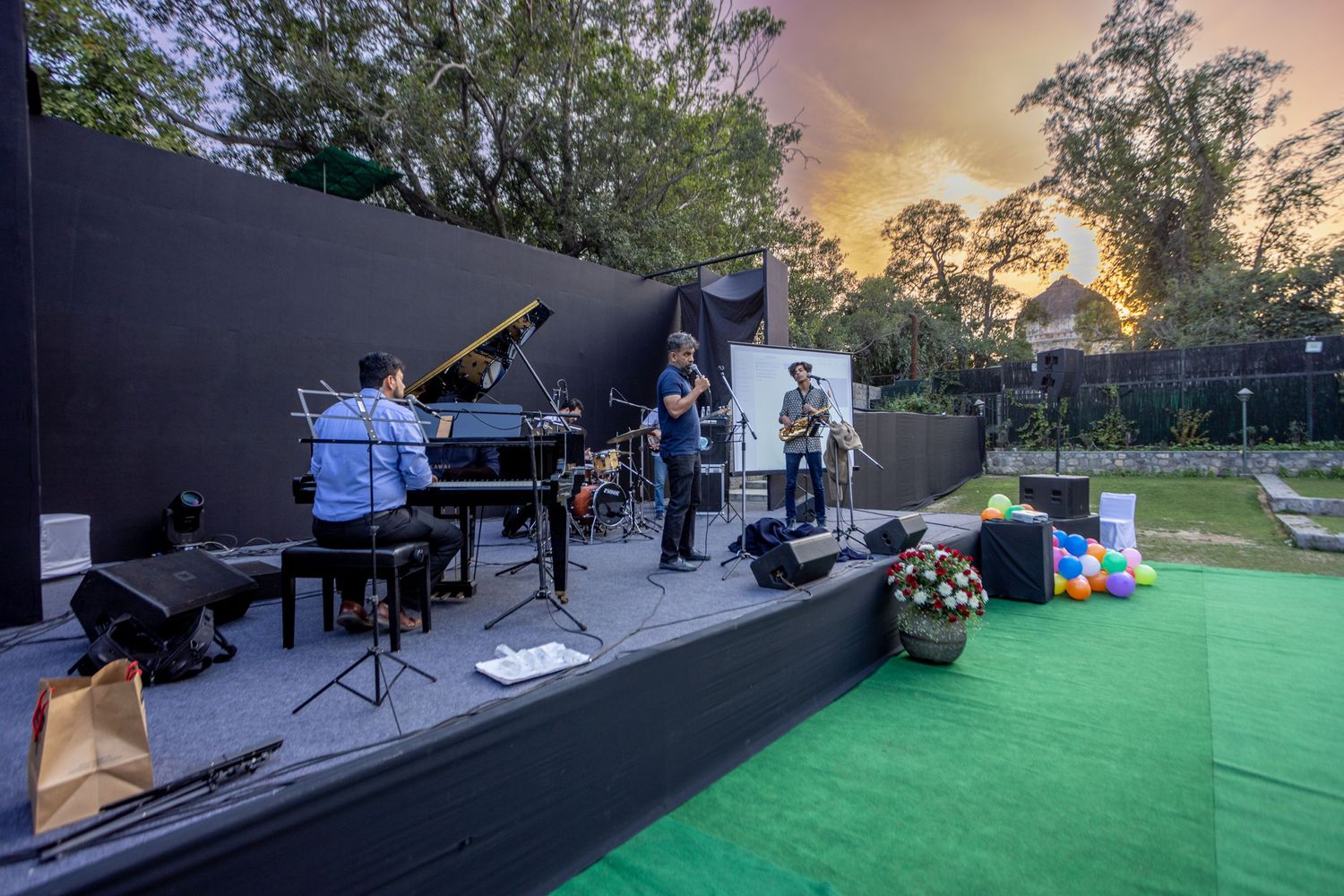 Musicians perform on an outdoor stage with piano and band setup during sunset with green turf in foreground.