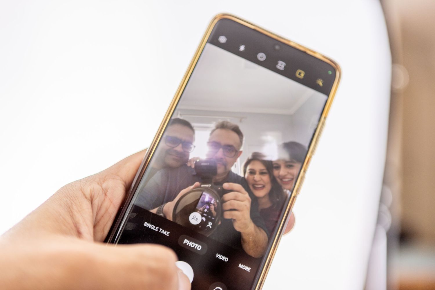Hand holding a smartphone displaying a camera app screen as a group of smiling people pose for a selfie photo.