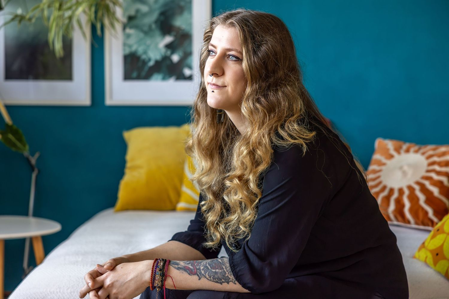 Woman with long wavy hair and tattoos sits on a white couch in a teal room with yellow and orange pillows.