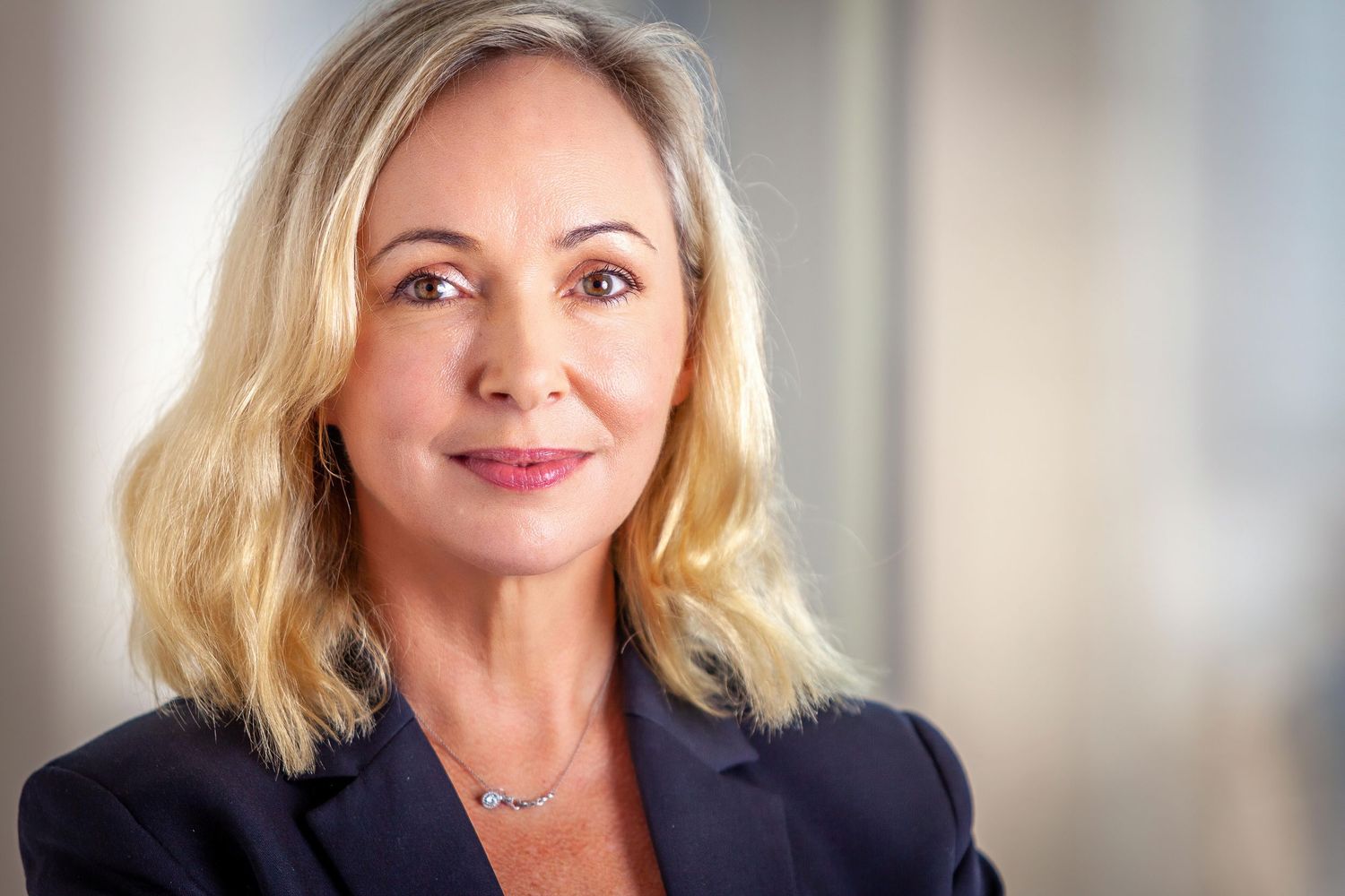 Professional headshot of a person in a dark blazer with blonde hair and a silver necklace.