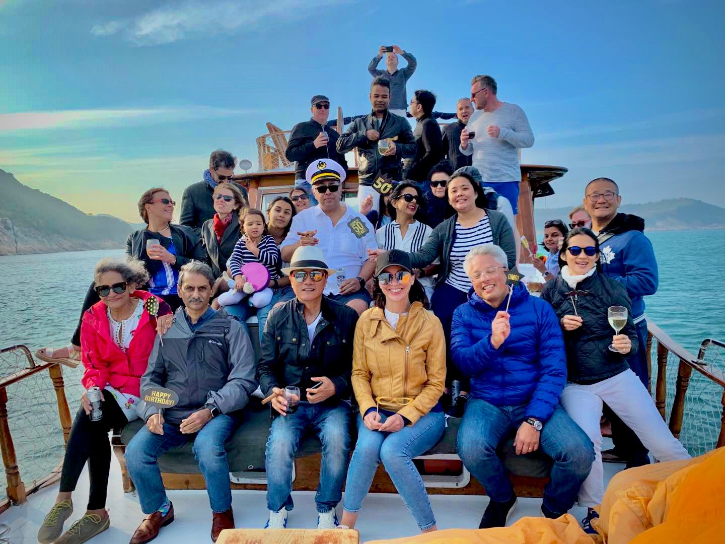 Large group gathered on boat deck with mountain and ocean backdrop.