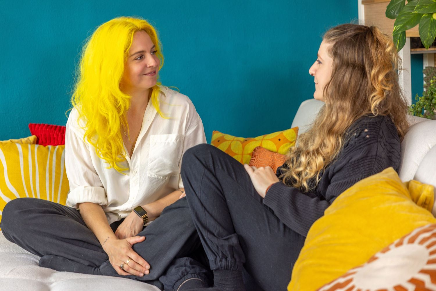 Two women sitting on a white couch chatting, one with bright yellow hair in a white shirt, the other in black, with colorful pillows.