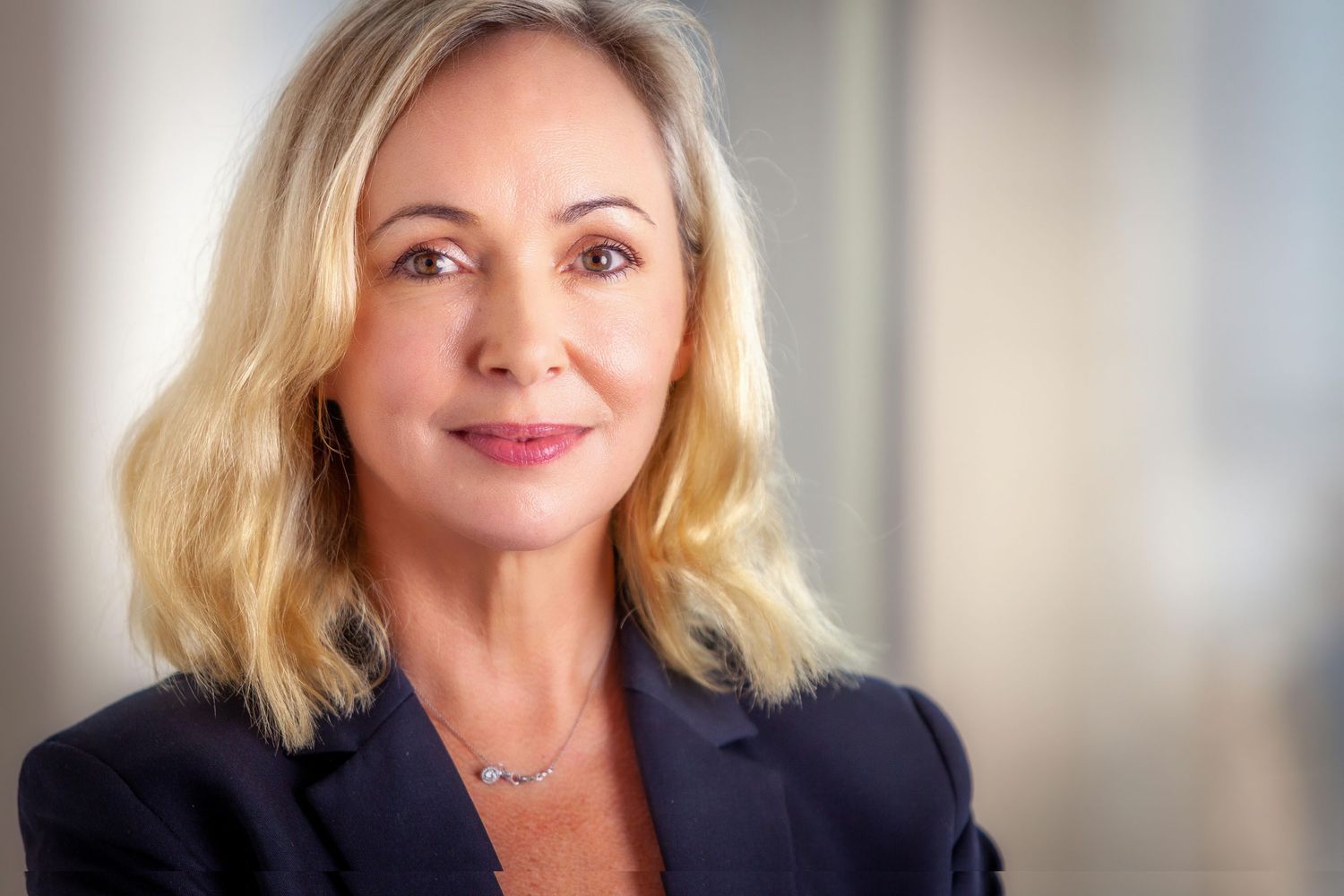 Professional headshot of a person with blonde hair wearing a navy blazer against a light background.