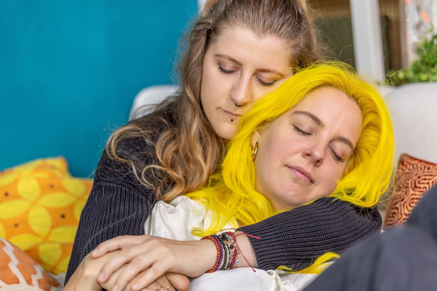 Two women sharing a tender embrace, one with yellow hair resting on the other's shoulder on a colorful sofa.